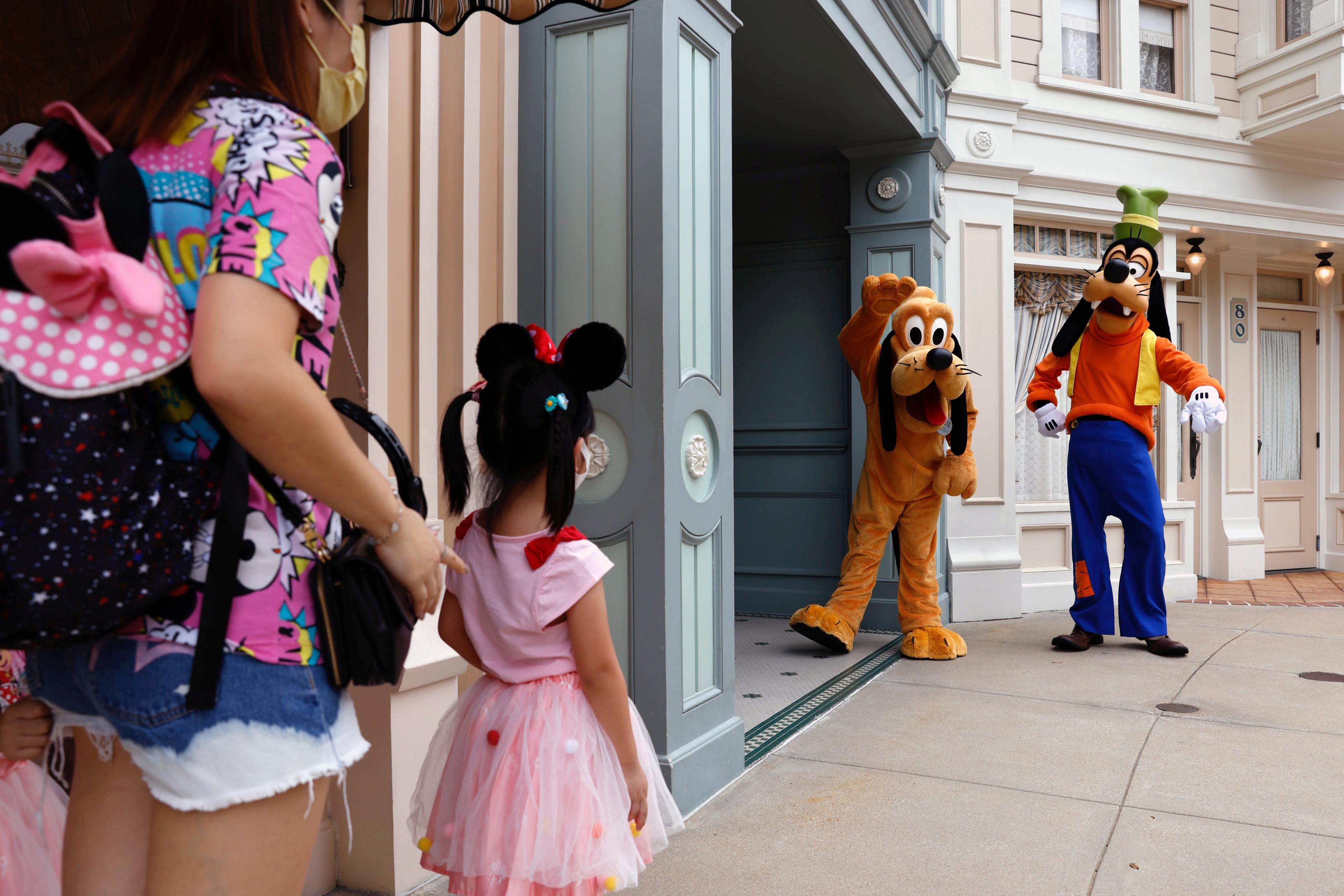 Photo of a child in a Minnie Mouse outfit meeting Pluto and Goofy characters at a theme park entrance with an adult nearby.