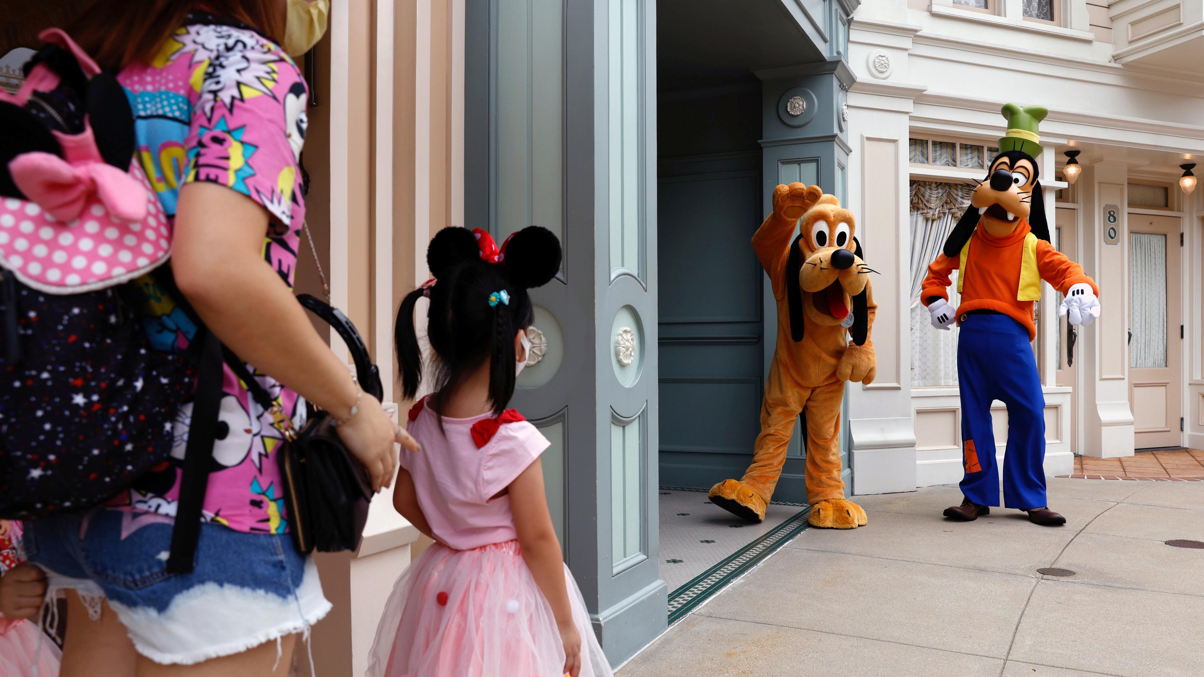 Photo of a child in a Minnie Mouse outfit meeting Pluto and Goofy characters at a theme park entrance with an adult nearby.