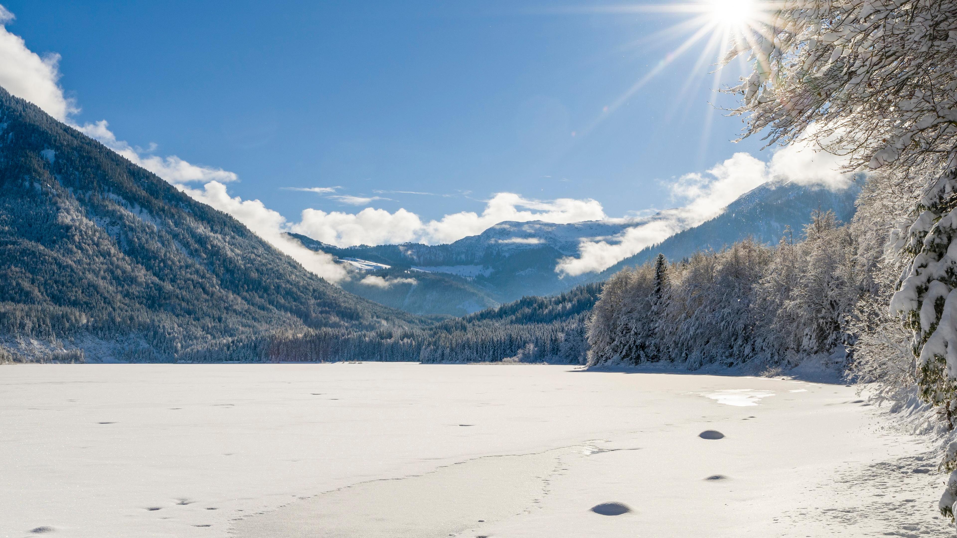 Photo of a snowy mountain landscape with a sunlit frozen lake, clear blue sky and snow-covered trees on the right.
