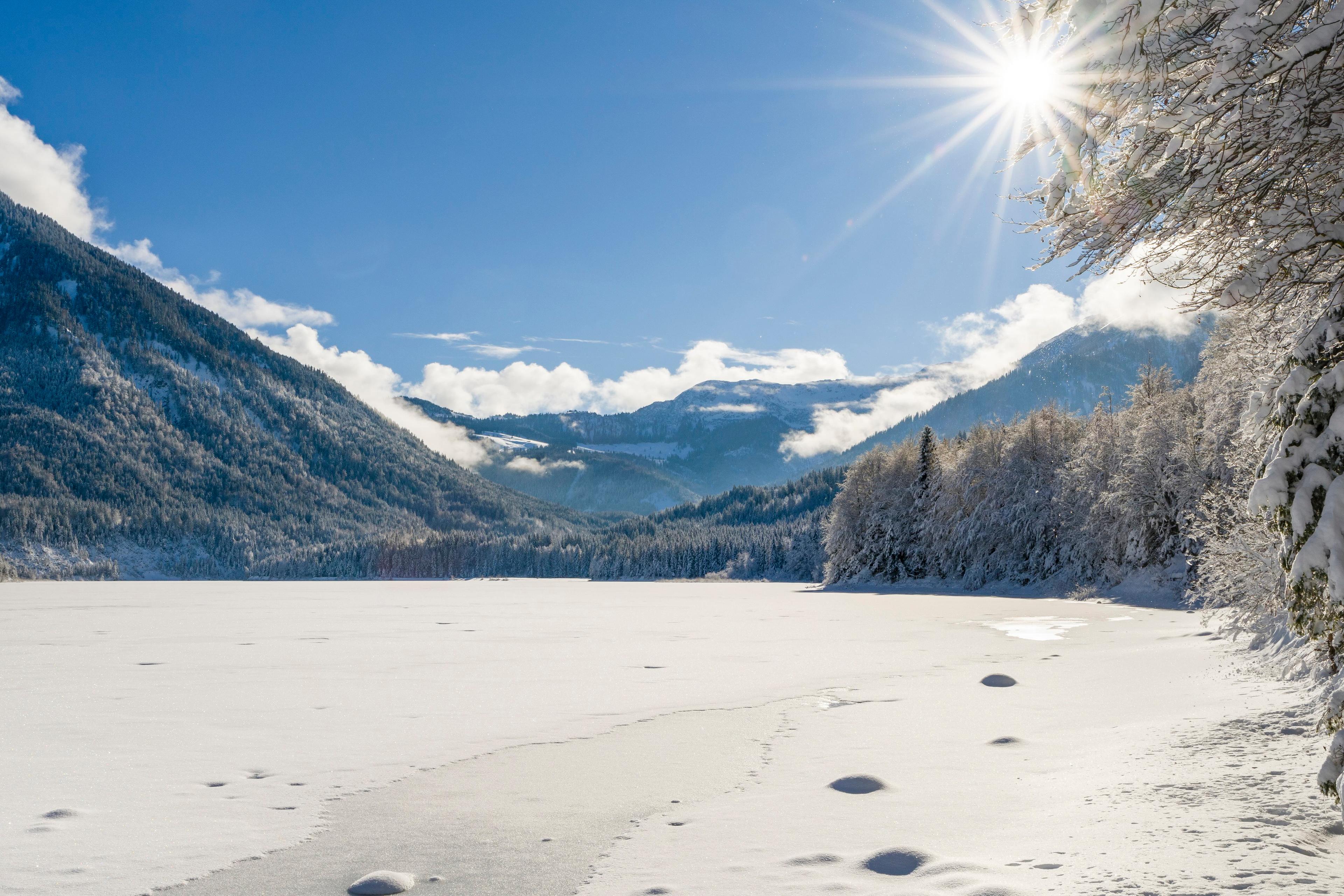 Photo of a snowy mountain landscape with a sunlit frozen lake, clear blue sky and snow-covered trees on the right.