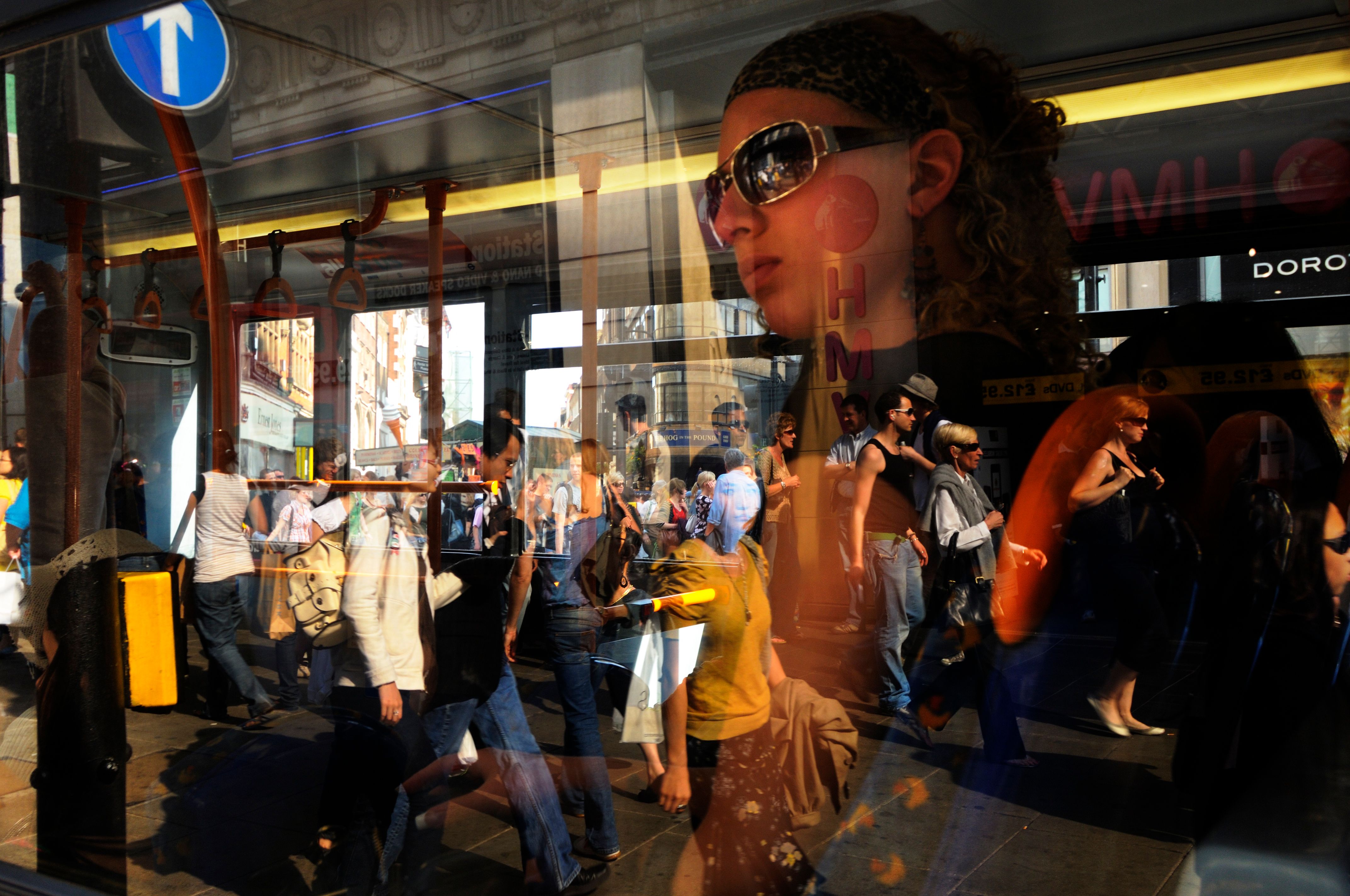 Photo of bus window reflection, showing people walking in a busy urban scene with a woman wearing sunglasses in focus.