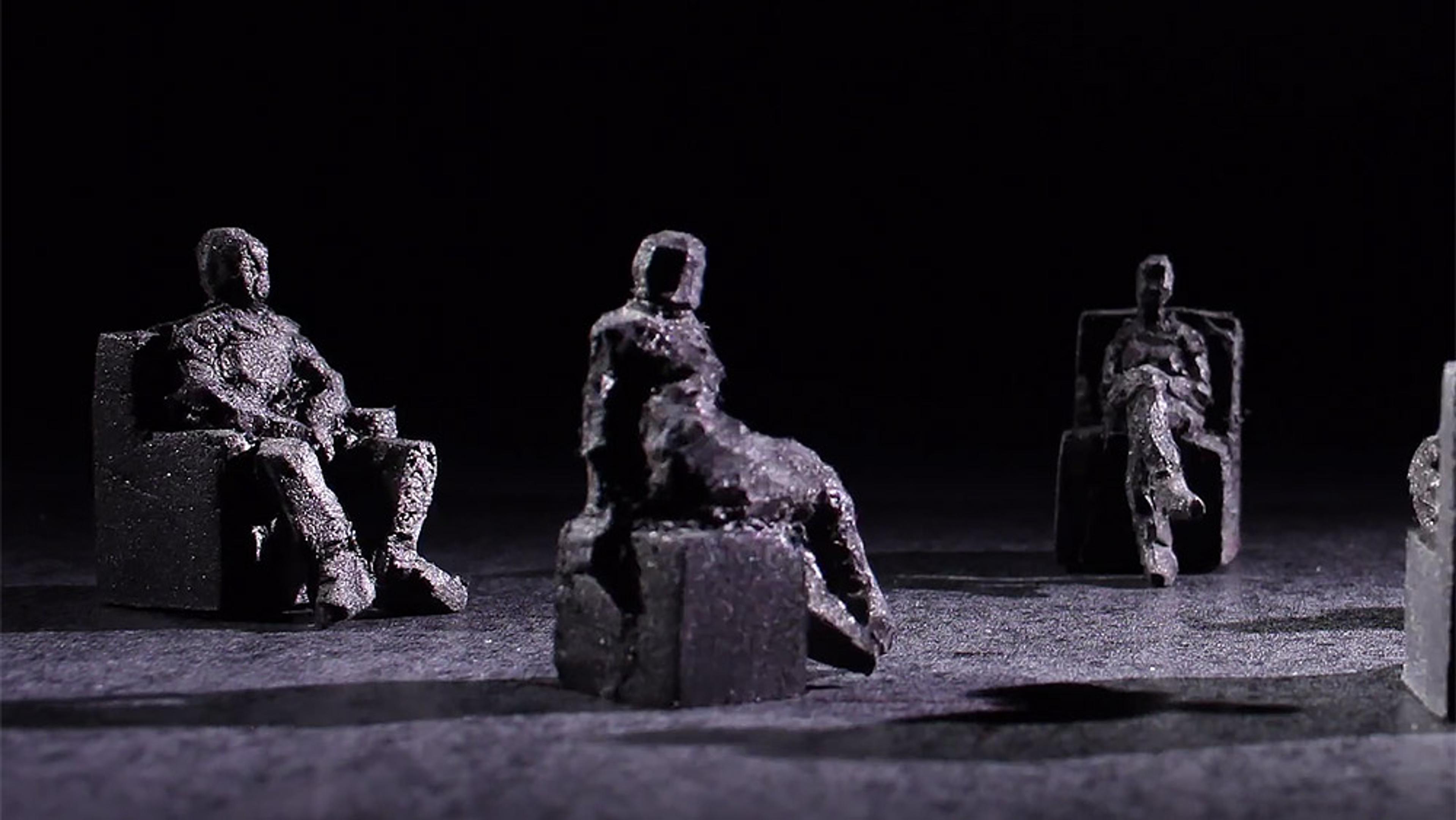 Photo of three dark, rough-textured clay sculptures of seated humans illuminated against a dark background.