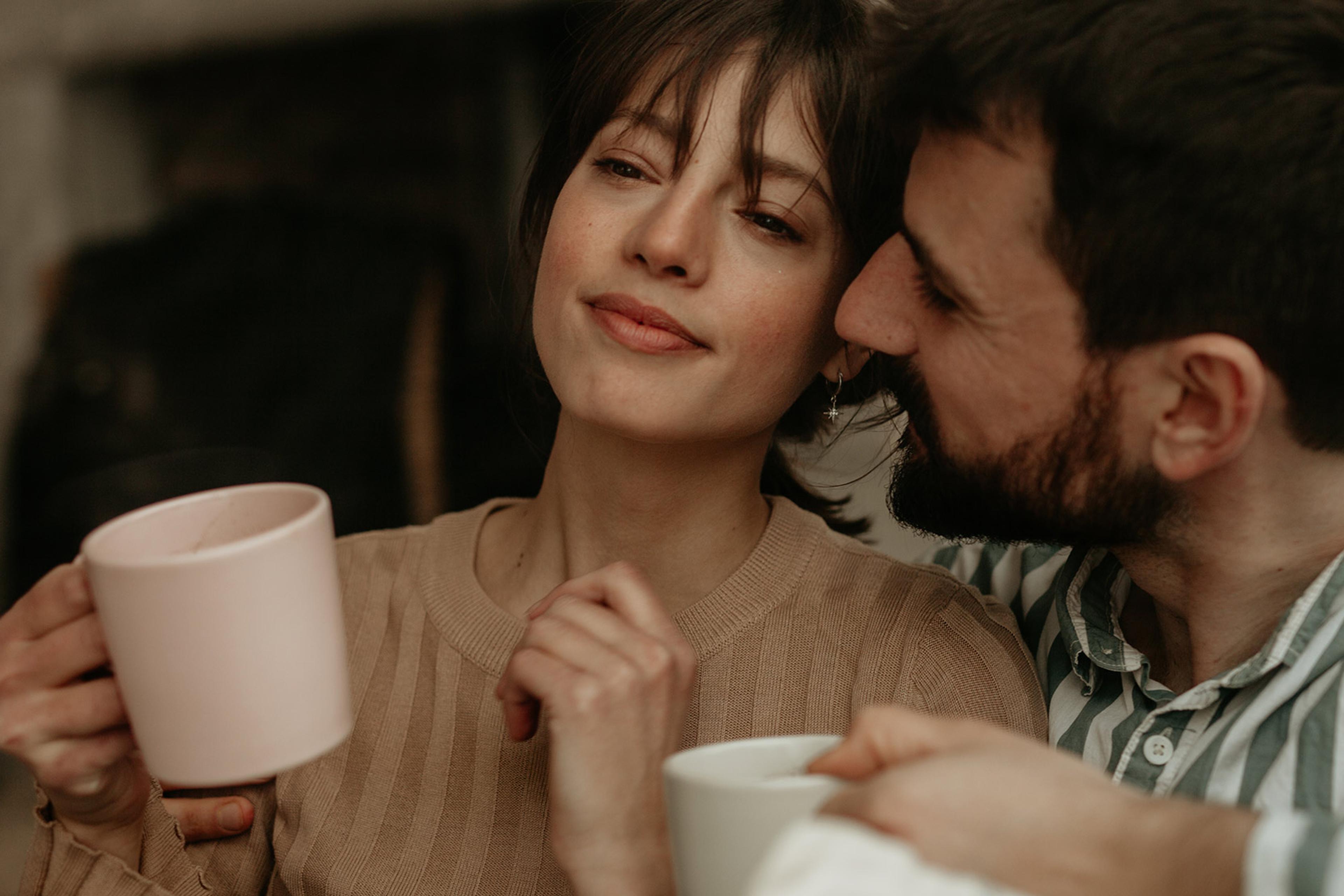 A couple enjoying a cosy moment, the woman holds a pink mug while the man, in a striped shirt, leans in closely, both smiling softly.