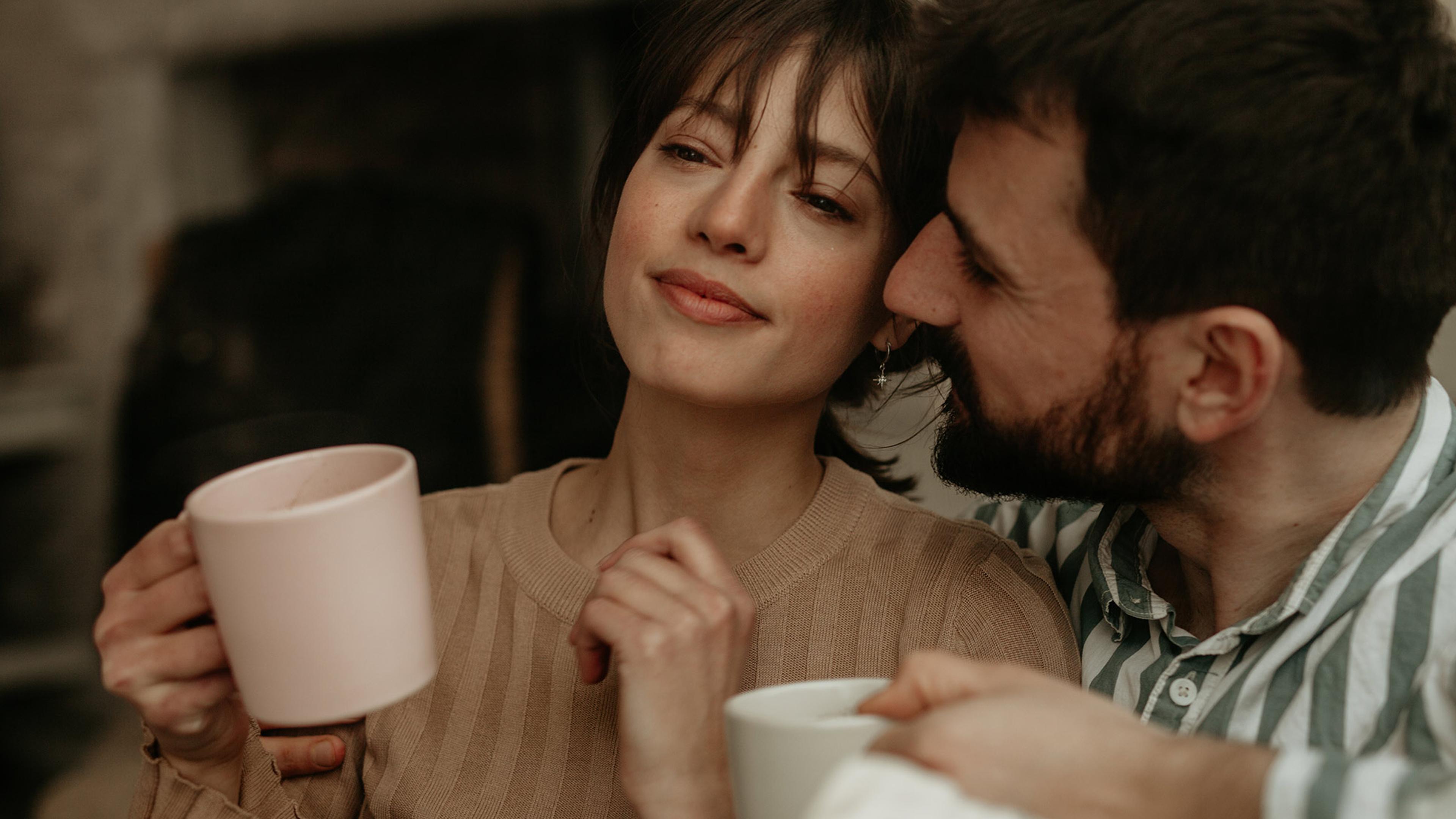A couple enjoying a cosy moment, the woman holds a pink mug while the man, in a striped shirt, leans in closely, both smiling softly.