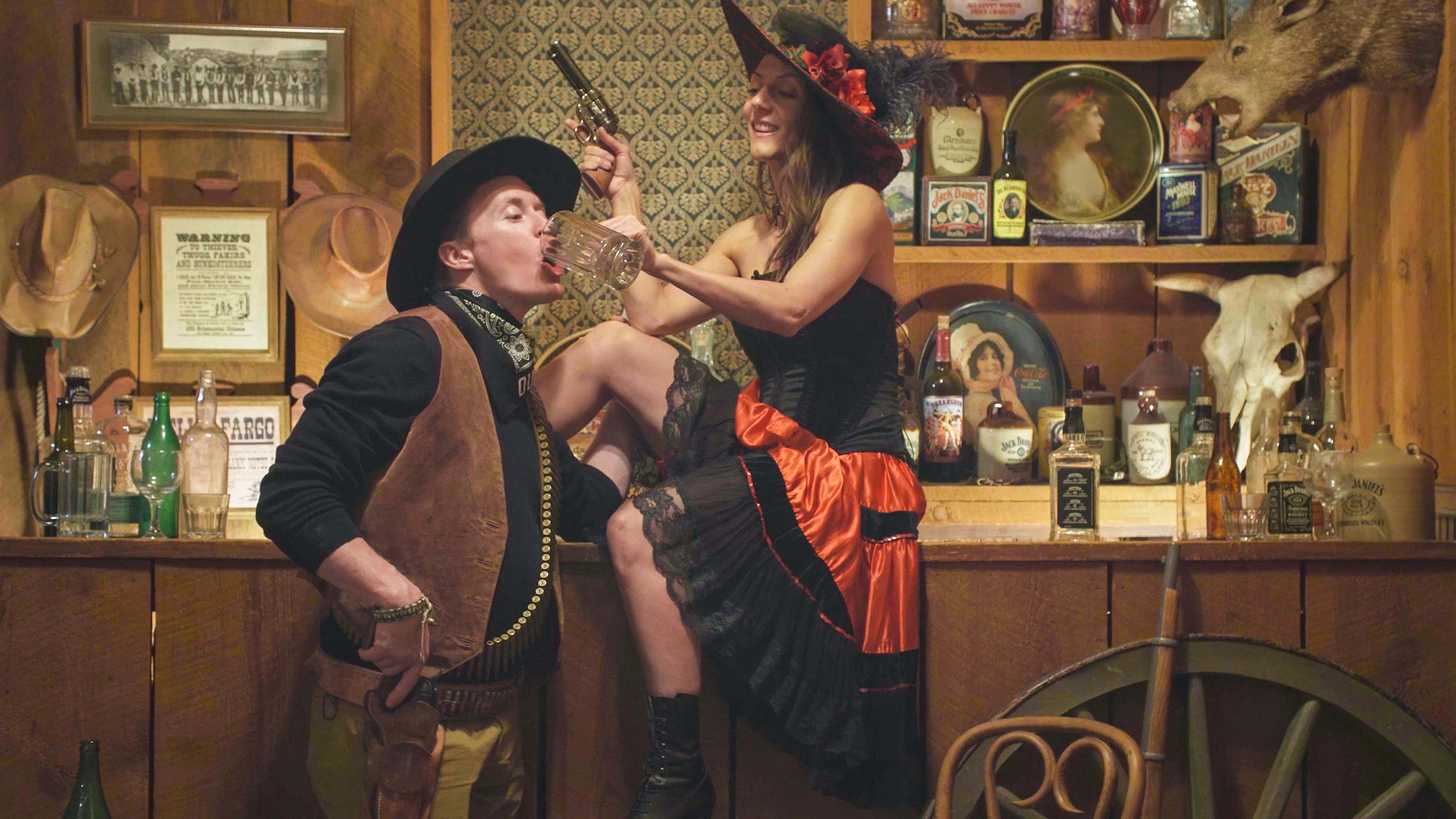 Vintage-style photo of a cowboy in a saloon drinking from a mug held by a saloon girl holding a gun at the bar.