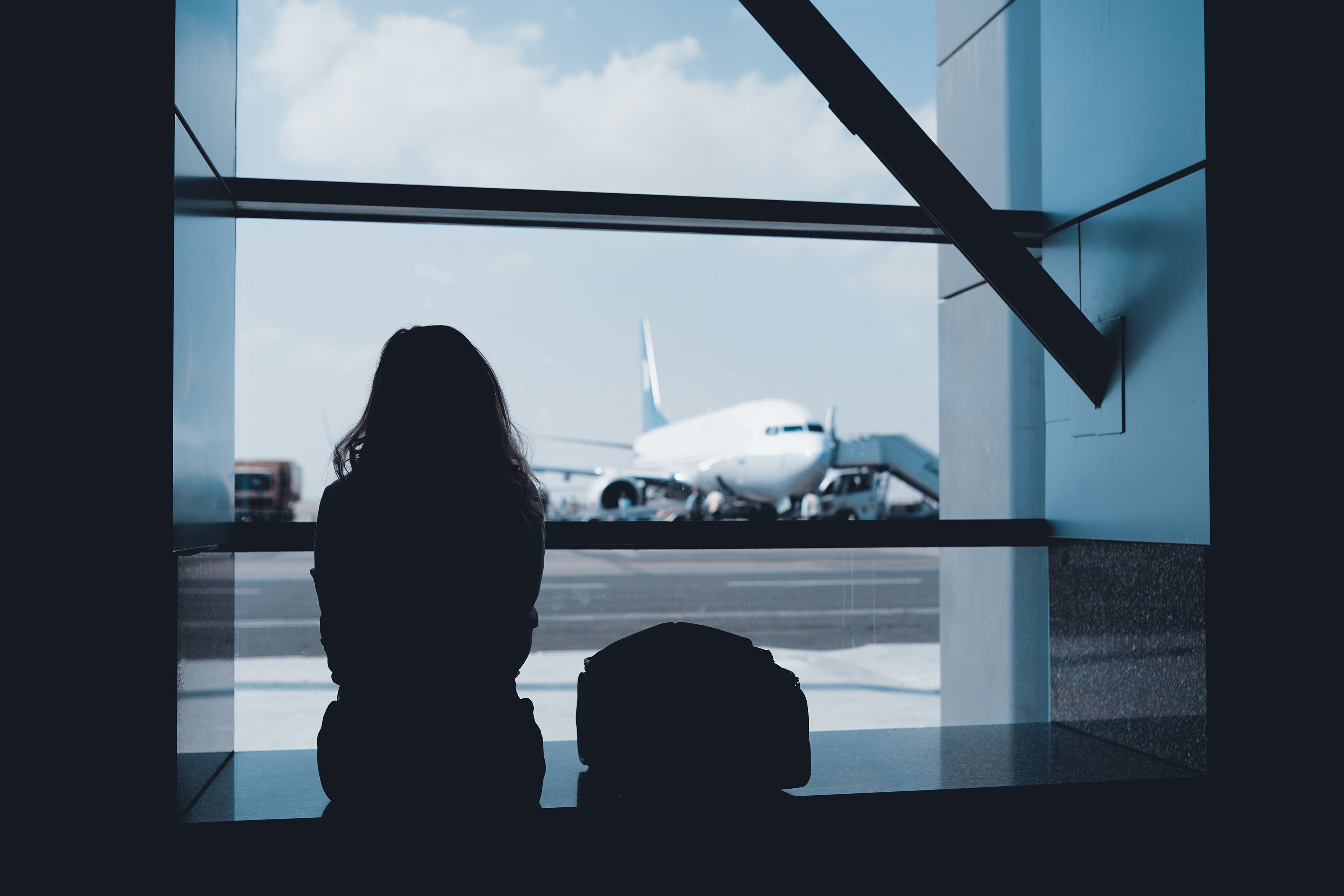 A silhouette of a person at an airport window watching a plane on the tarmac under a blue sky.