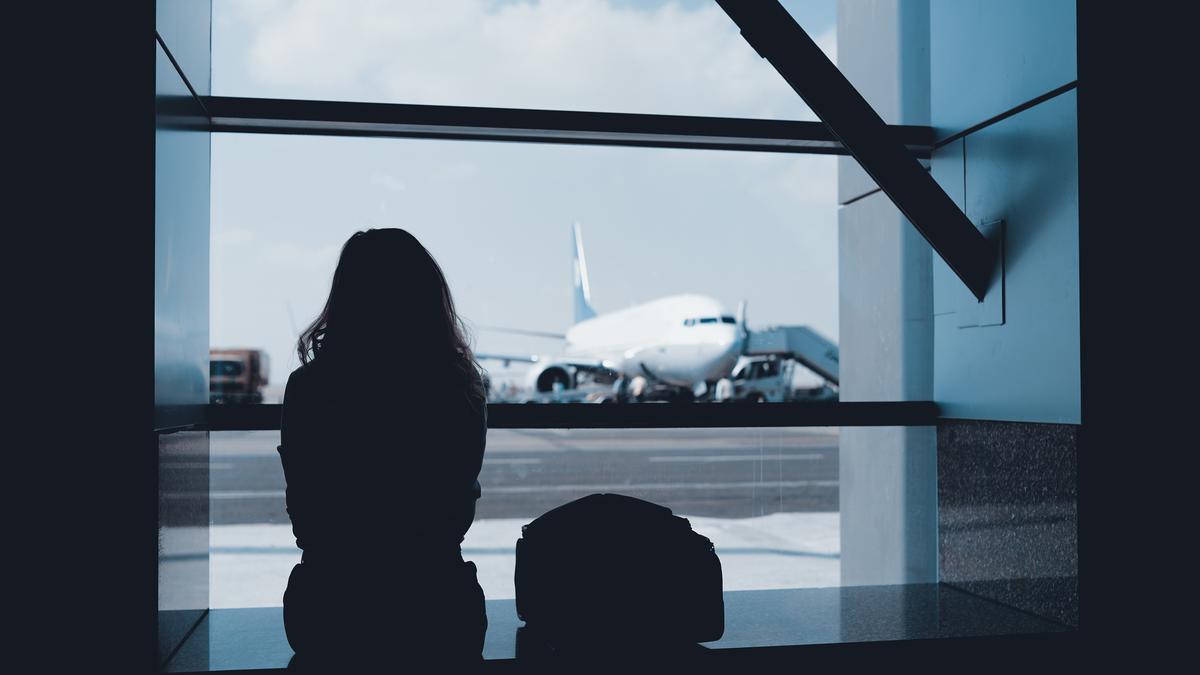 A silhouette of a person at an airport window watching a plane on the tarmac under a blue sky.
