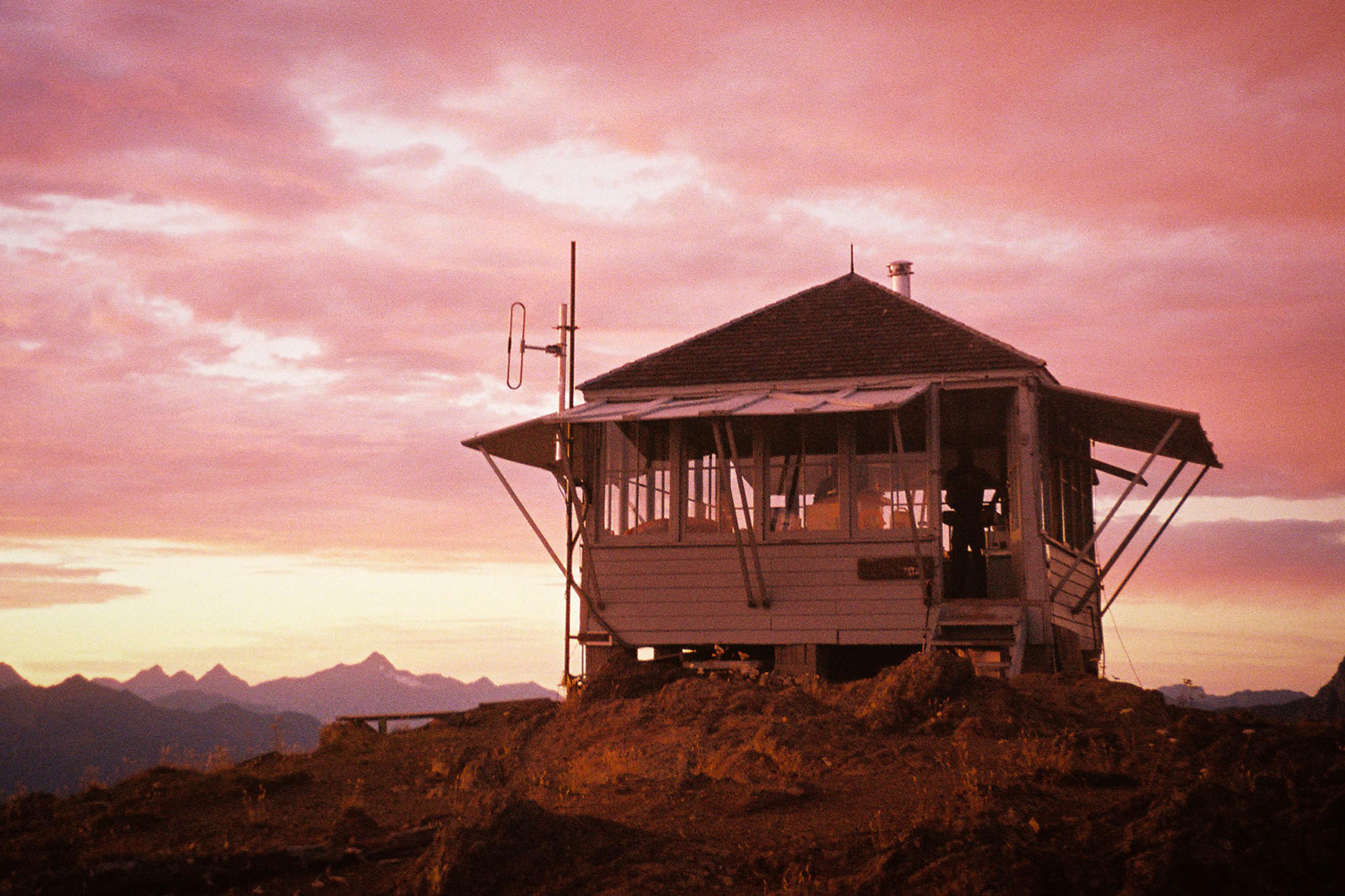 A small wooden cabin on a hill at sunset with a pink sky and mountains in the background.