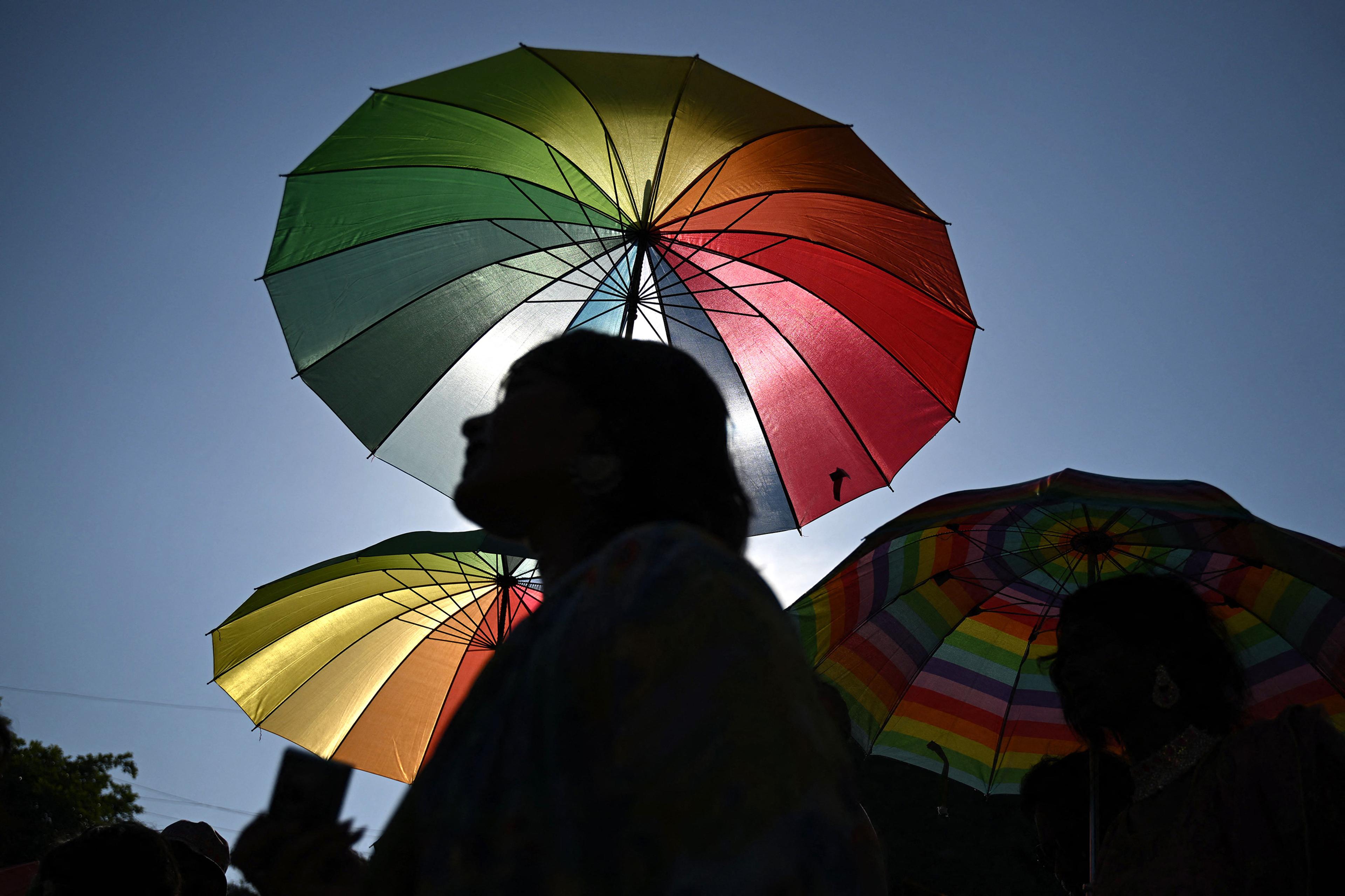 Silhouetted people under large vibrant, colourful umbrellas against a clear blue sky.