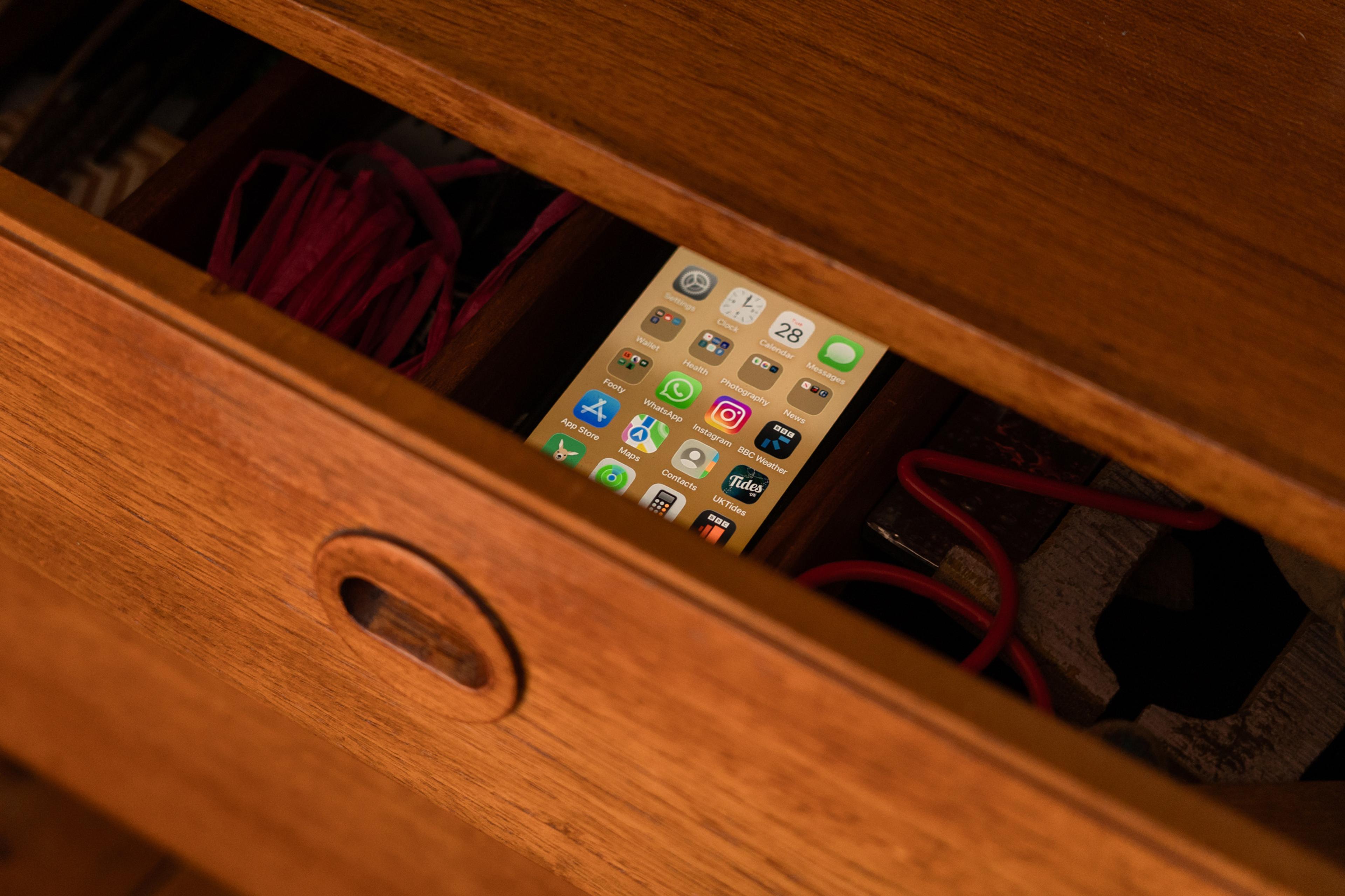 A smartphone with app icons visible inside an open wooden drawer with red cables partially visible.