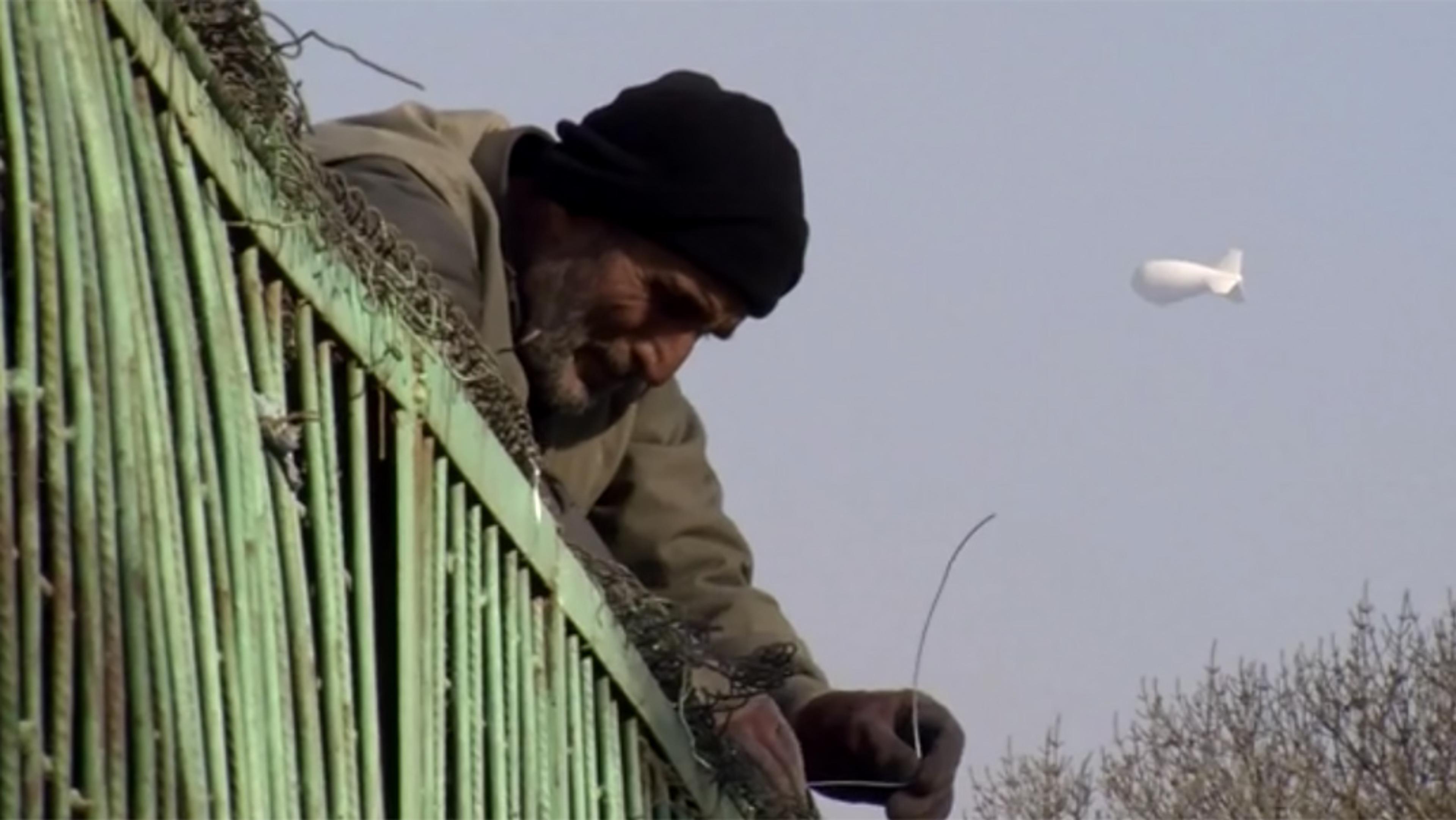 A man in a beanie fixing a green chain-link fence with a white blimp hovering in the sky in the background.