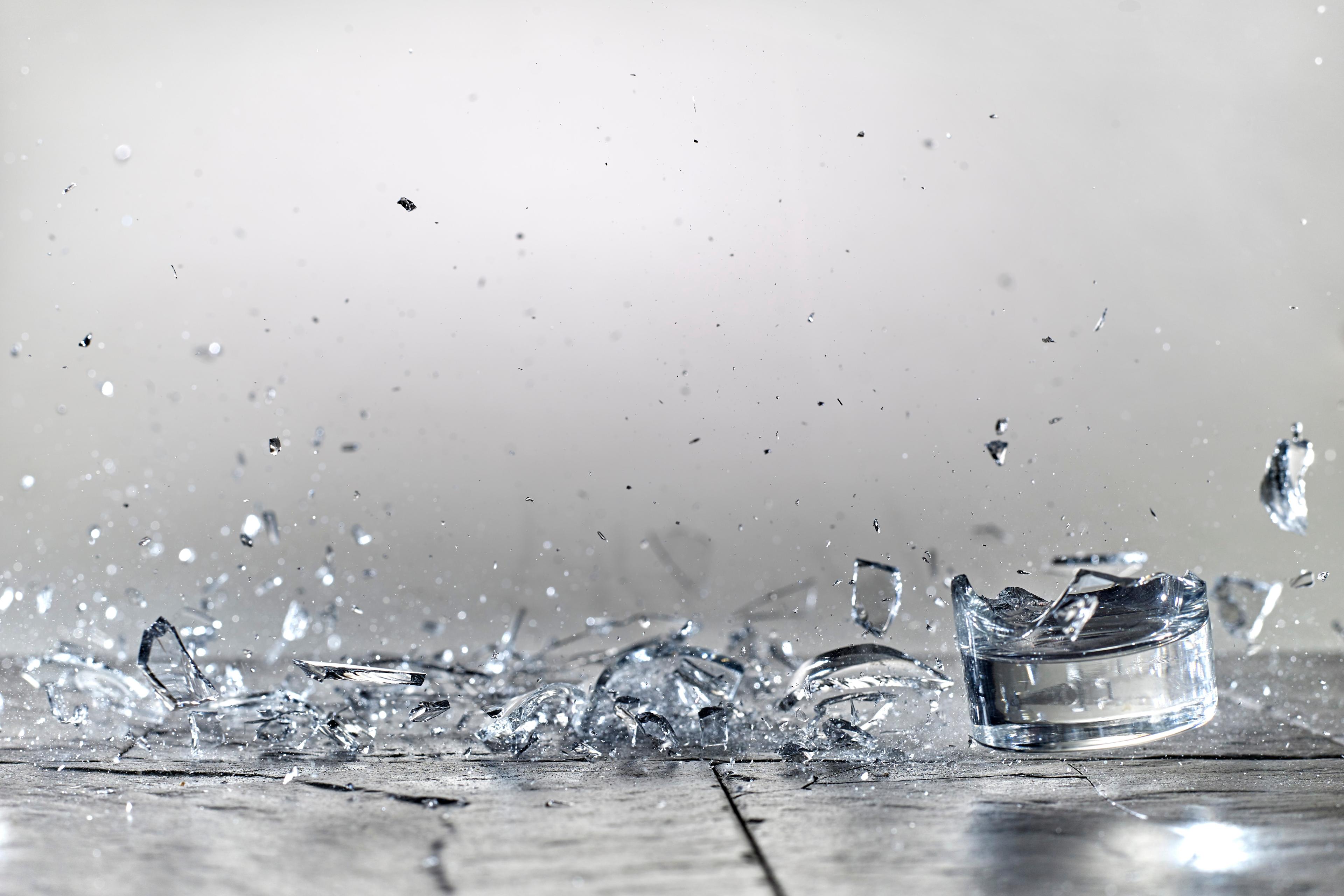 Photo of a glass shattering on a grey surface with shards scattered mid-air and on the ground.