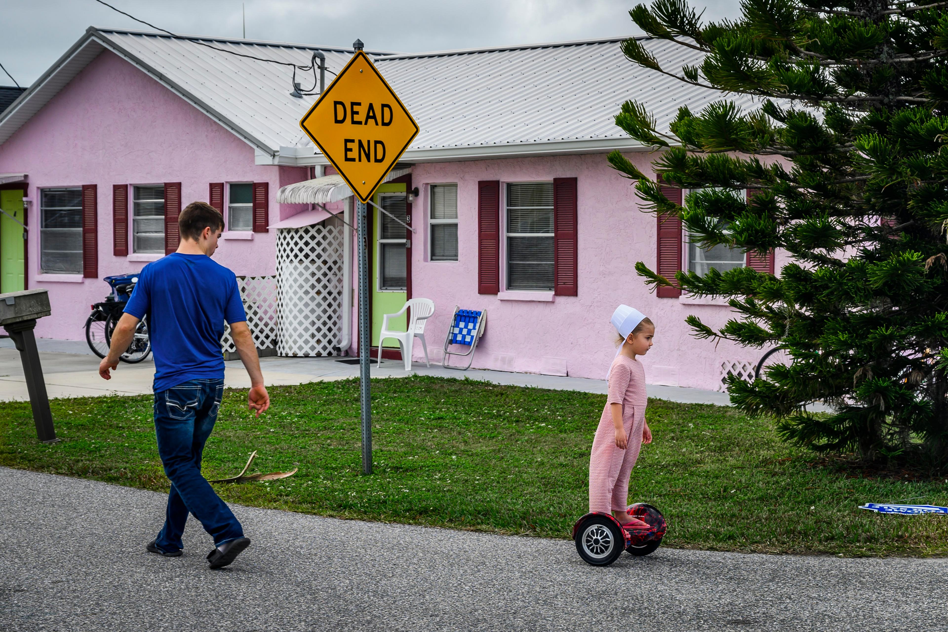 Photo of a man walking and a child on a hoverboard near a pink house and a ‘Dead End’ sign on a suburban street.