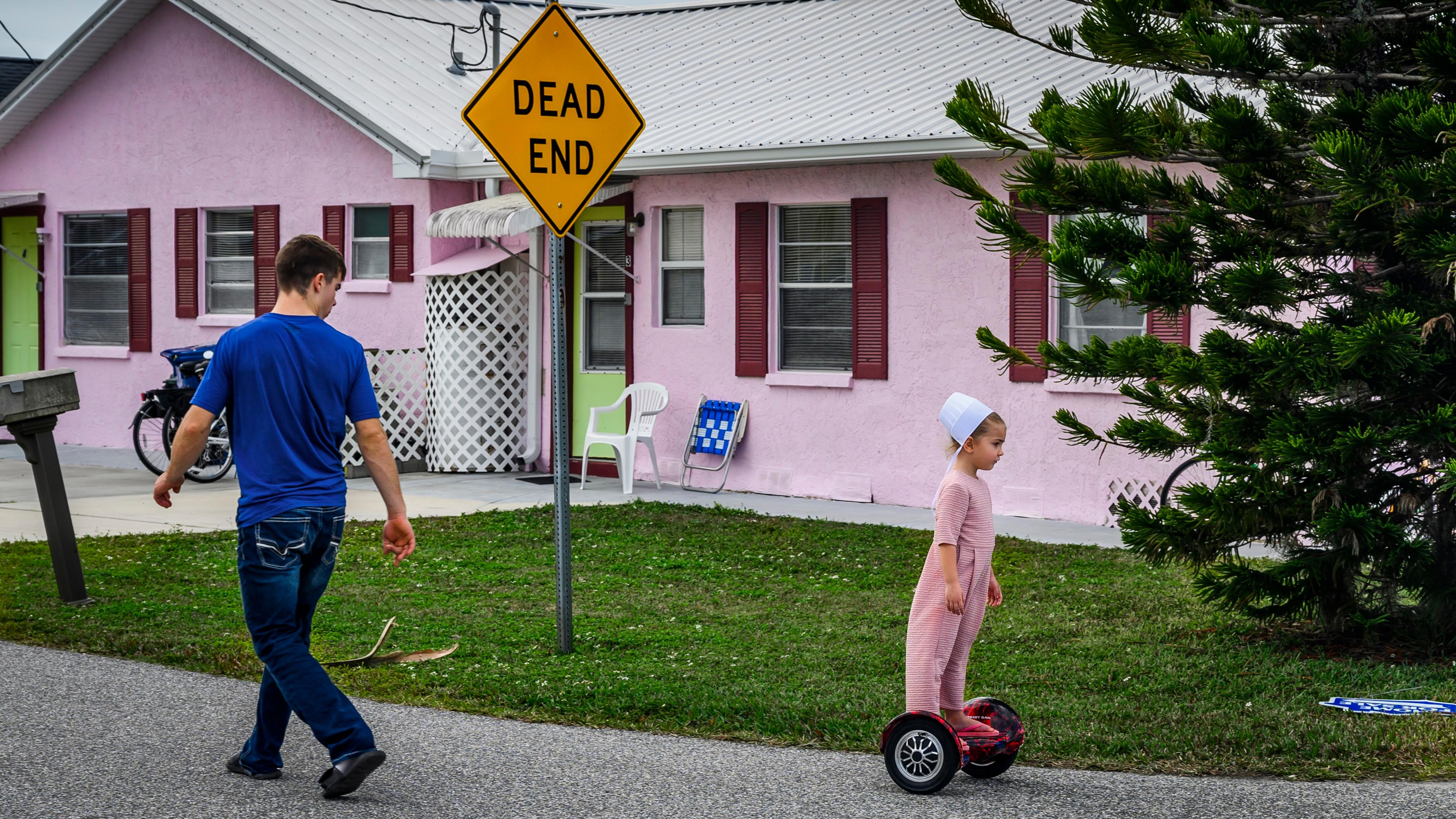 Photo of a man walking and a child on a hoverboard near a pink house and a ‘Dead End’ sign on a suburban street.