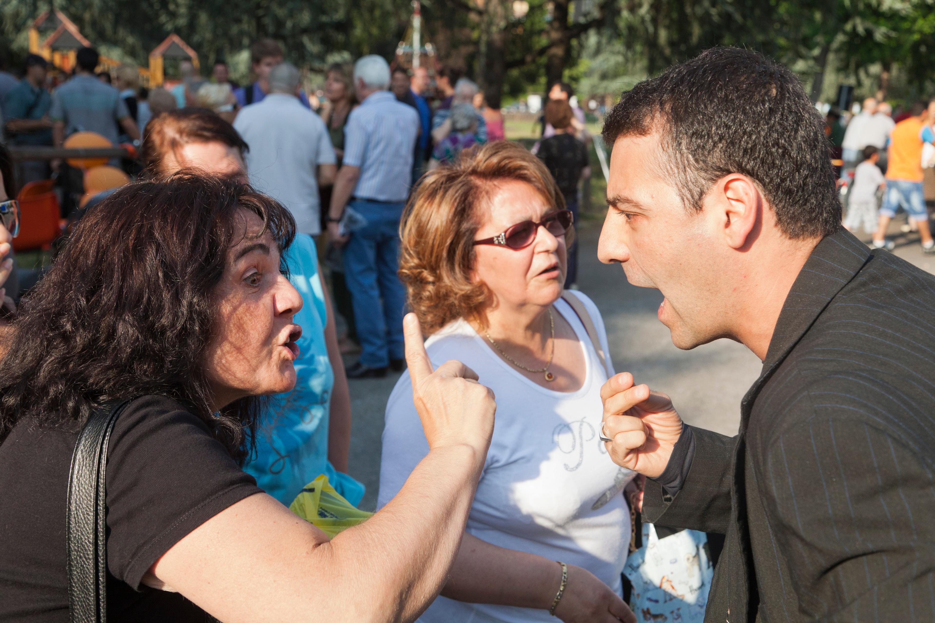 Photo of two women and a man arguing animatedly in a crowded outdoor setting with trees in the background.