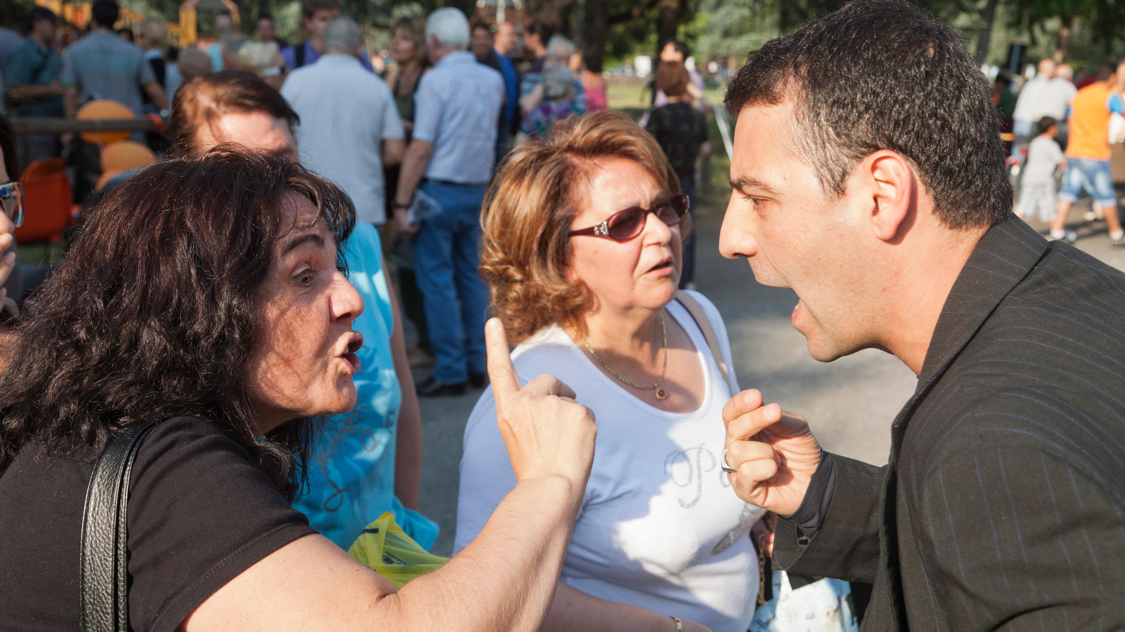 Photo of two women and a man arguing animatedly in a crowded outdoor setting with trees in the background.