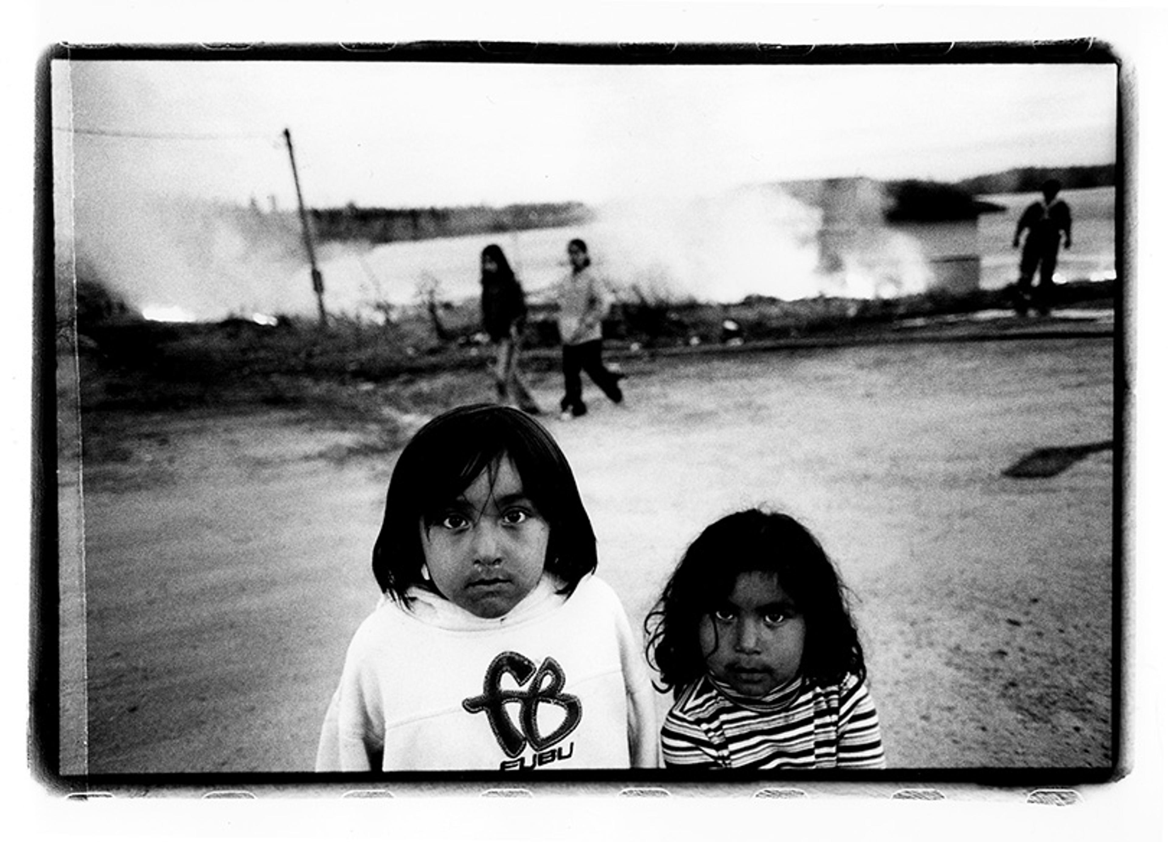 Black-and-white photo of two children standing outdoors with people walking behind and mist rising from a nearby body of water.