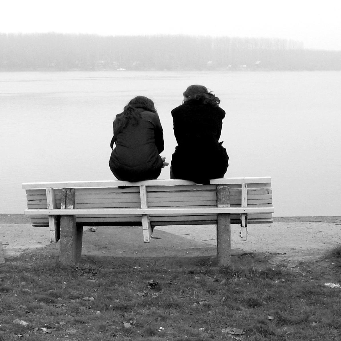 Black and white photo of two people sitting on a bench by a lake between two trees.