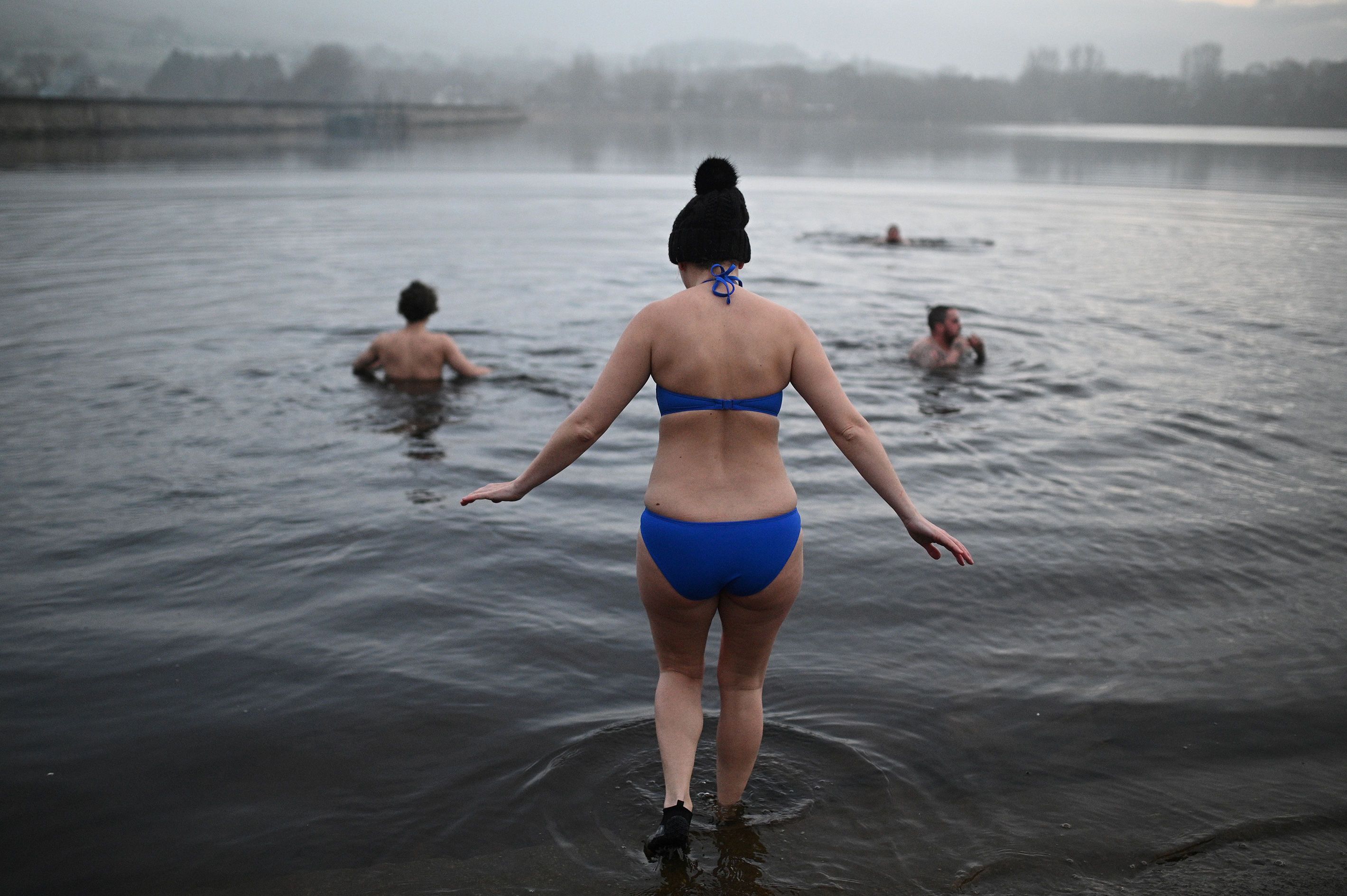 Photo of people swimming in a misty lake, with a person in a blue bikini entering the water in the foreground.