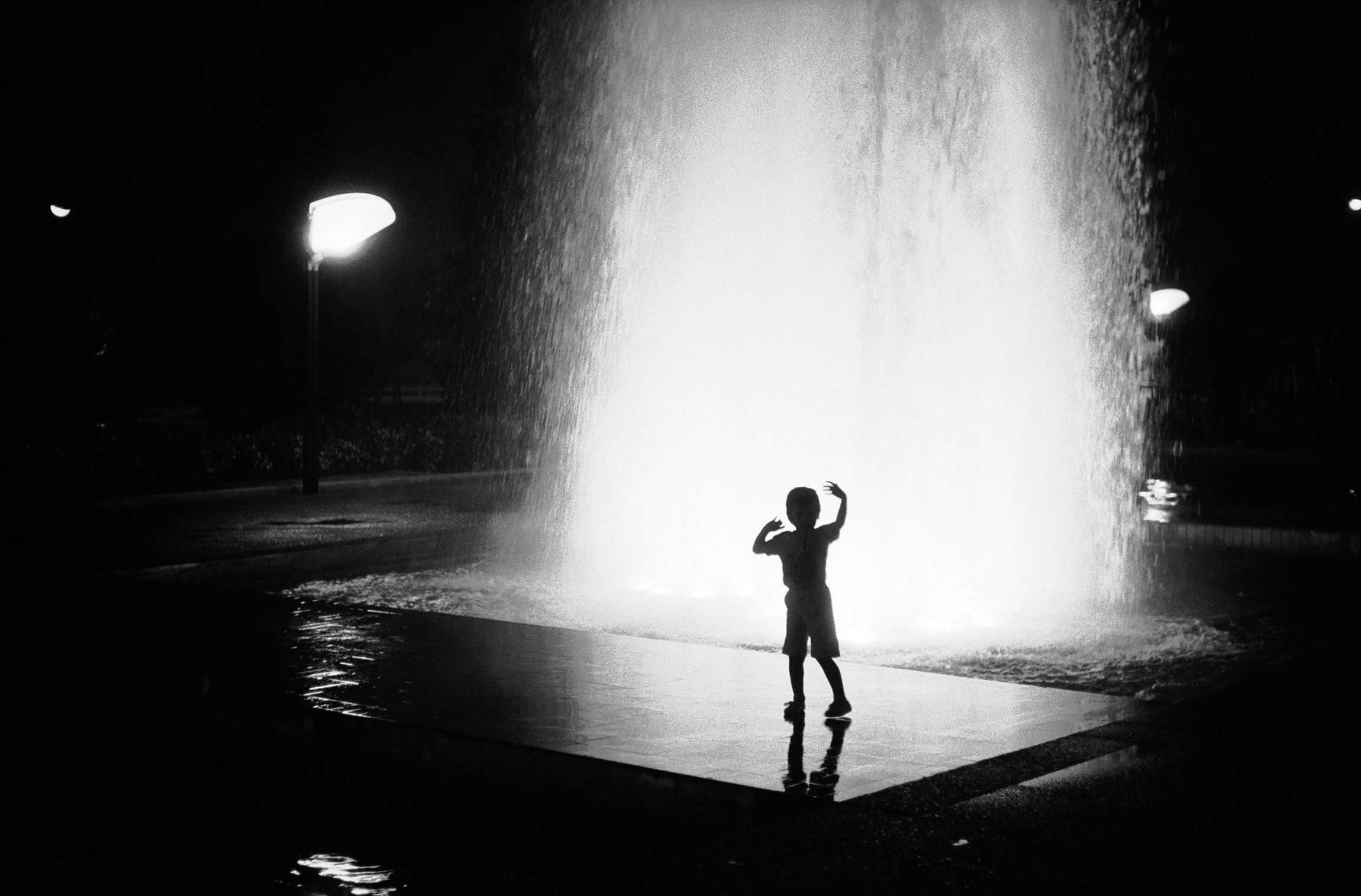 Black and white photo of a child silhouetted against a brightly lit fountain at night.