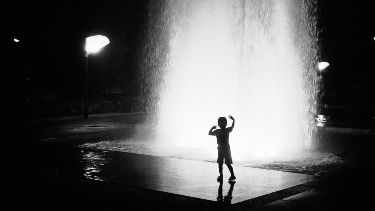 Black and white photo of a child silhouetted against a brightly lit fountain at night.
