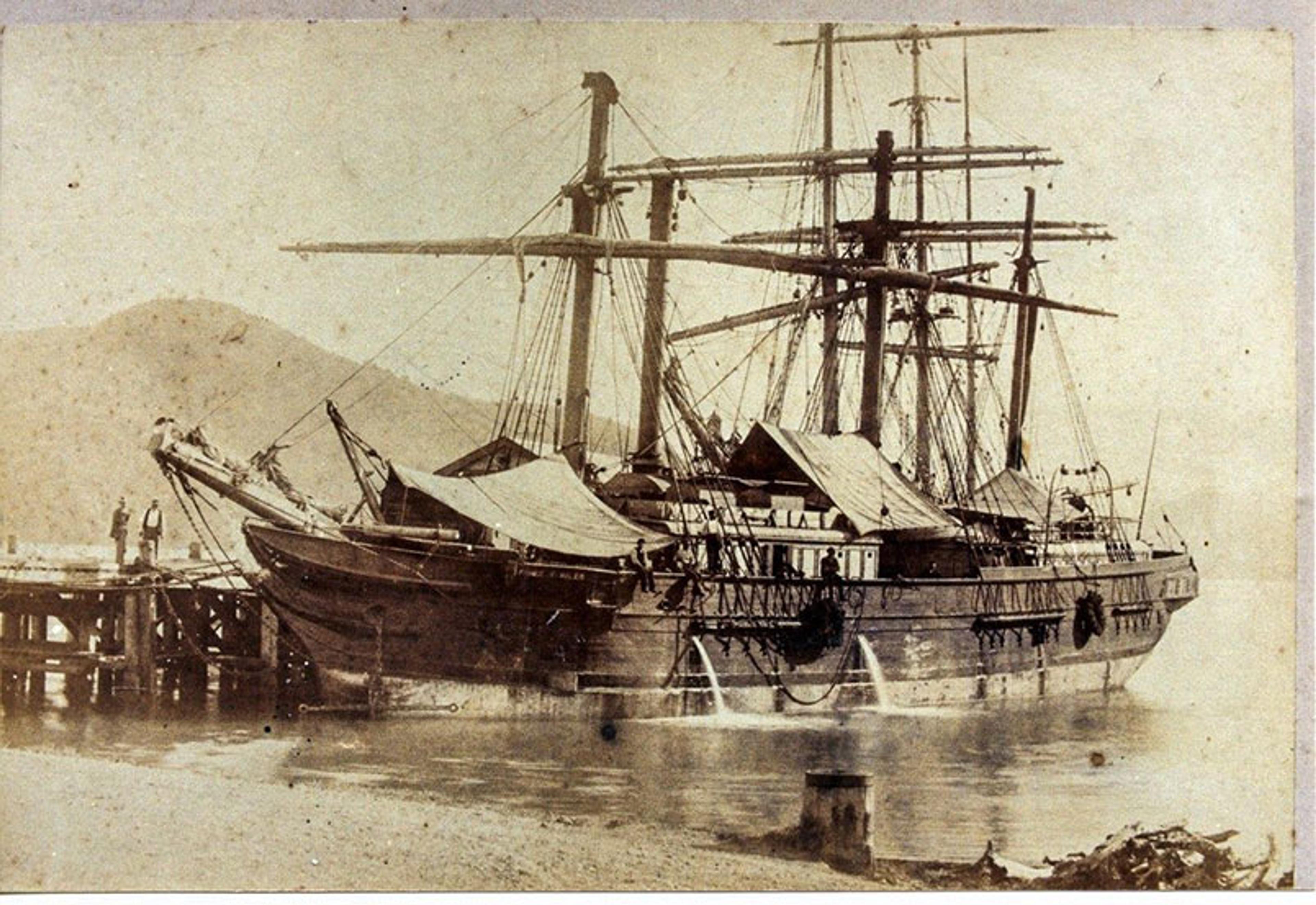 A sepia-toned photograph of an old sailing ship docked at a pier with mountains in the background and several sailors on board.