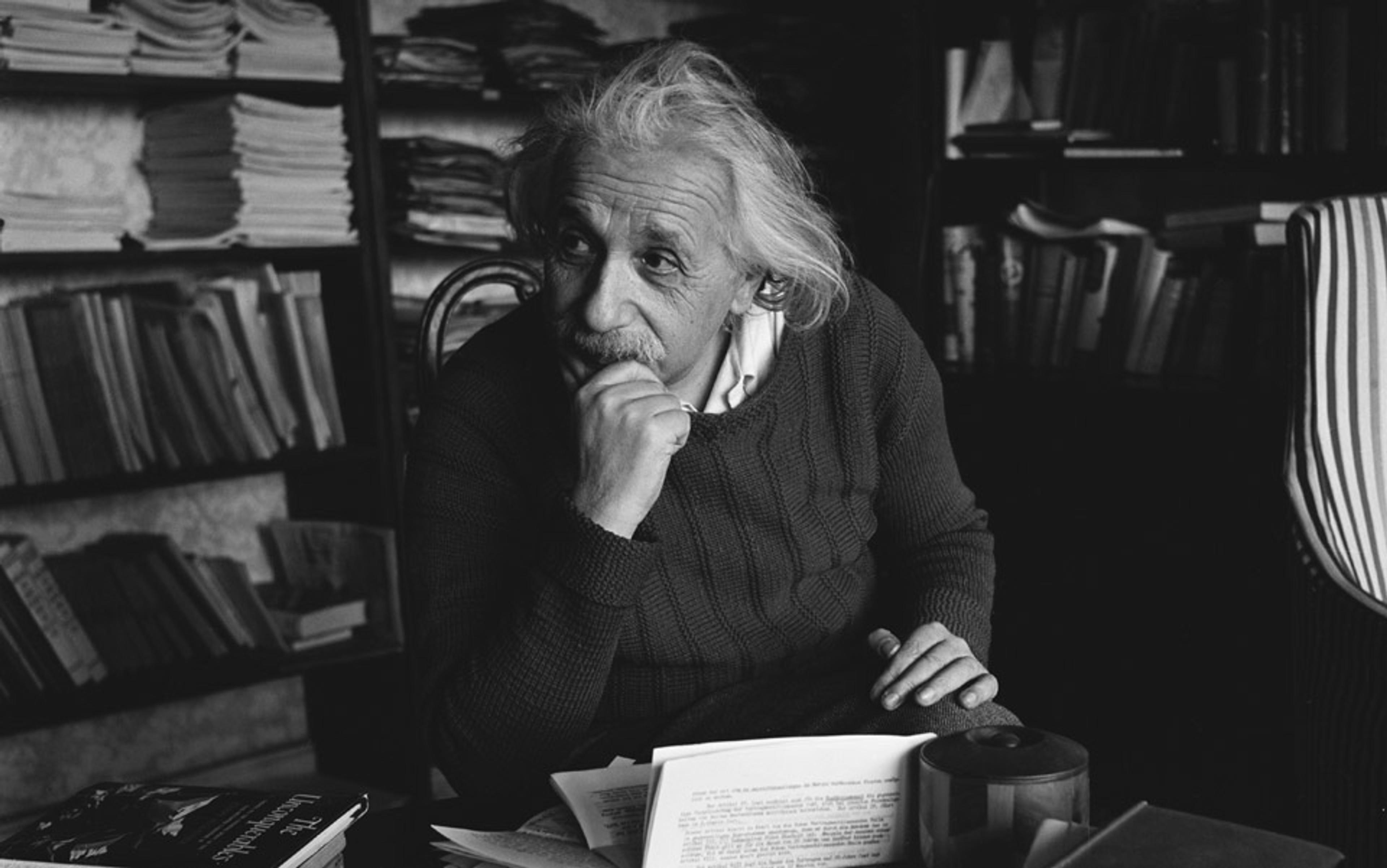 Black and white photo of Albert Einstein seated at a desk full of papers with bookshelves behind him.