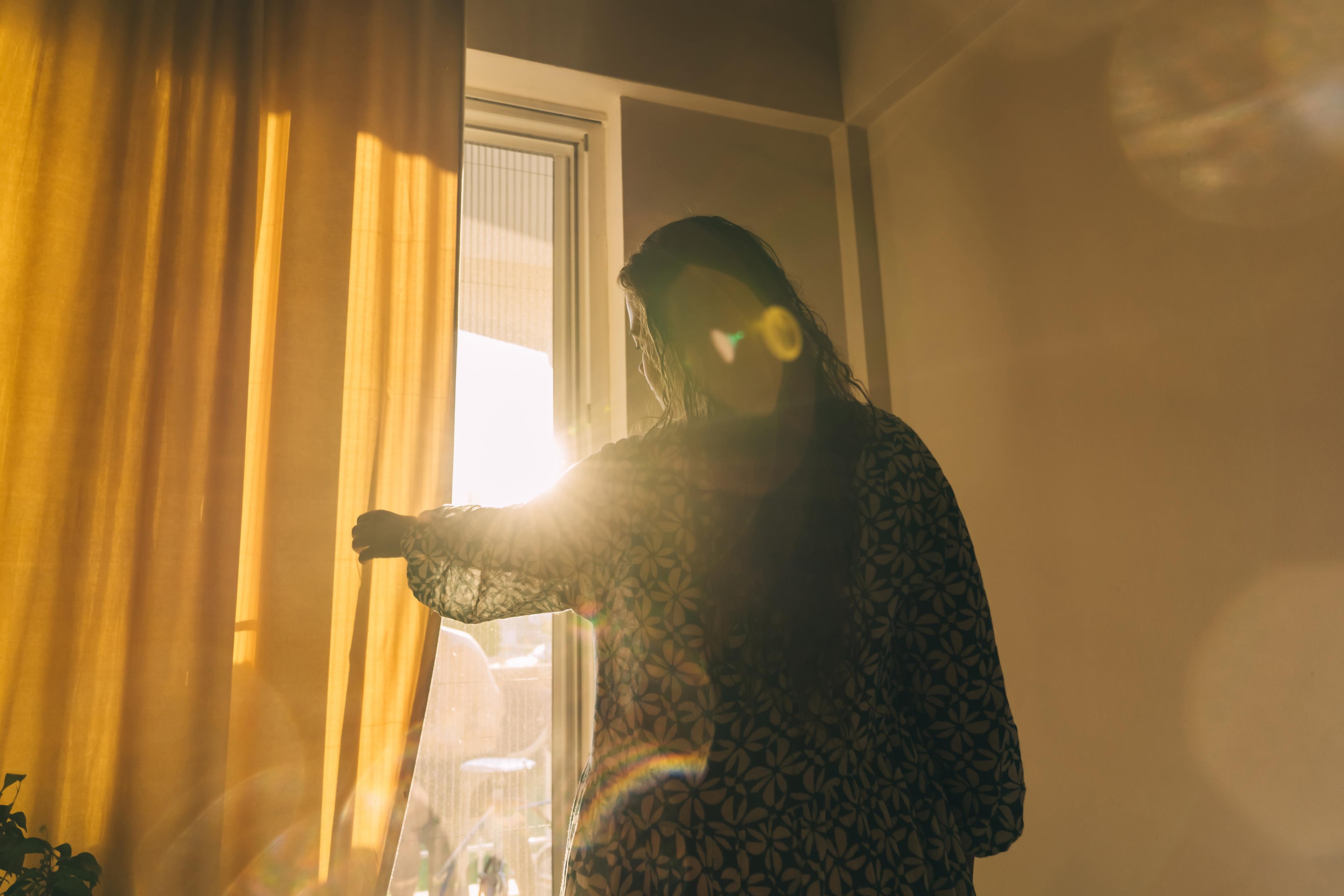A person opening yellow curtains with sunlight streaming in through a window, creating a warm, glowing effect.