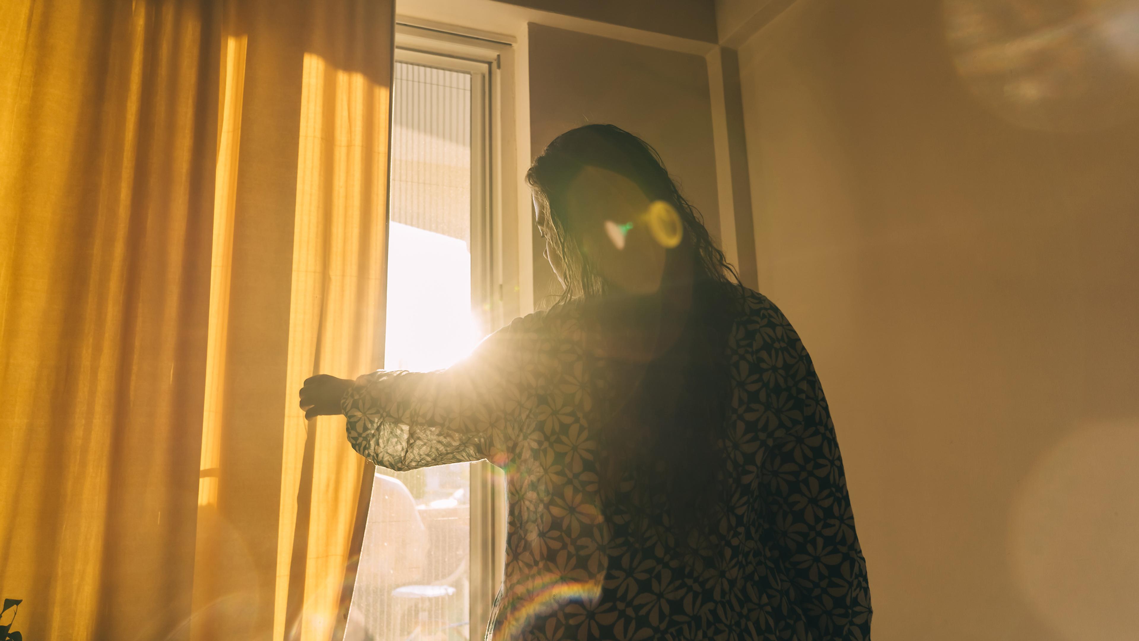 A person opening yellow curtains with sunlight streaming in through a window, creating a warm, glowing effect.