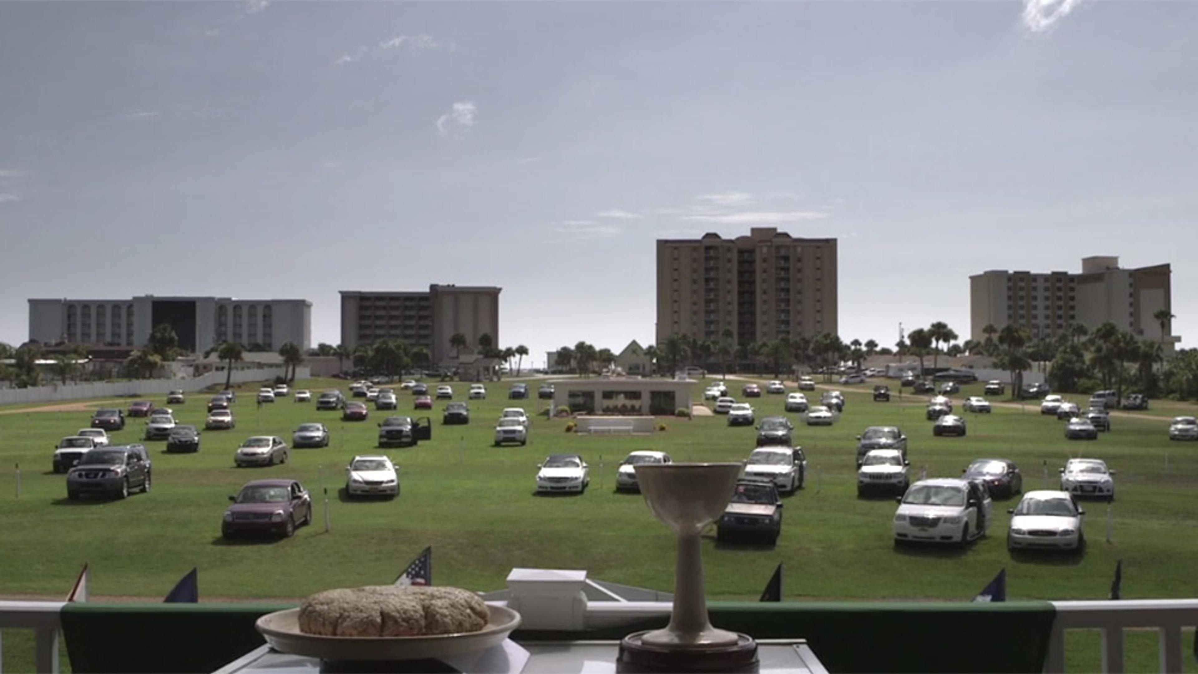 A drive-in church service with cars parked on a grass field, buildings in the background and communion elements in the foreground.