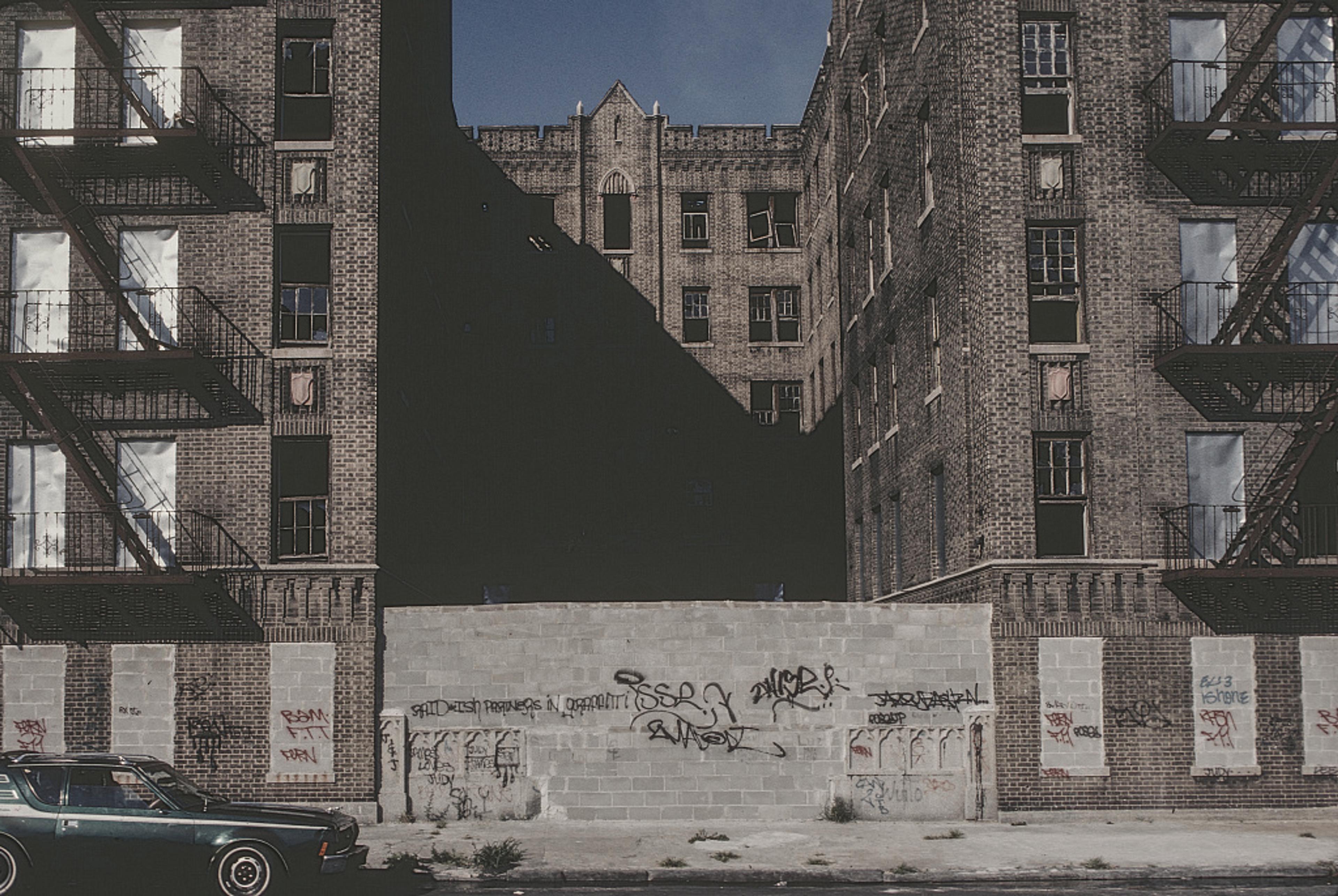 A cityscape with graffiti on brick buildings, an old car parked in front, and metal fire escapes on either side.