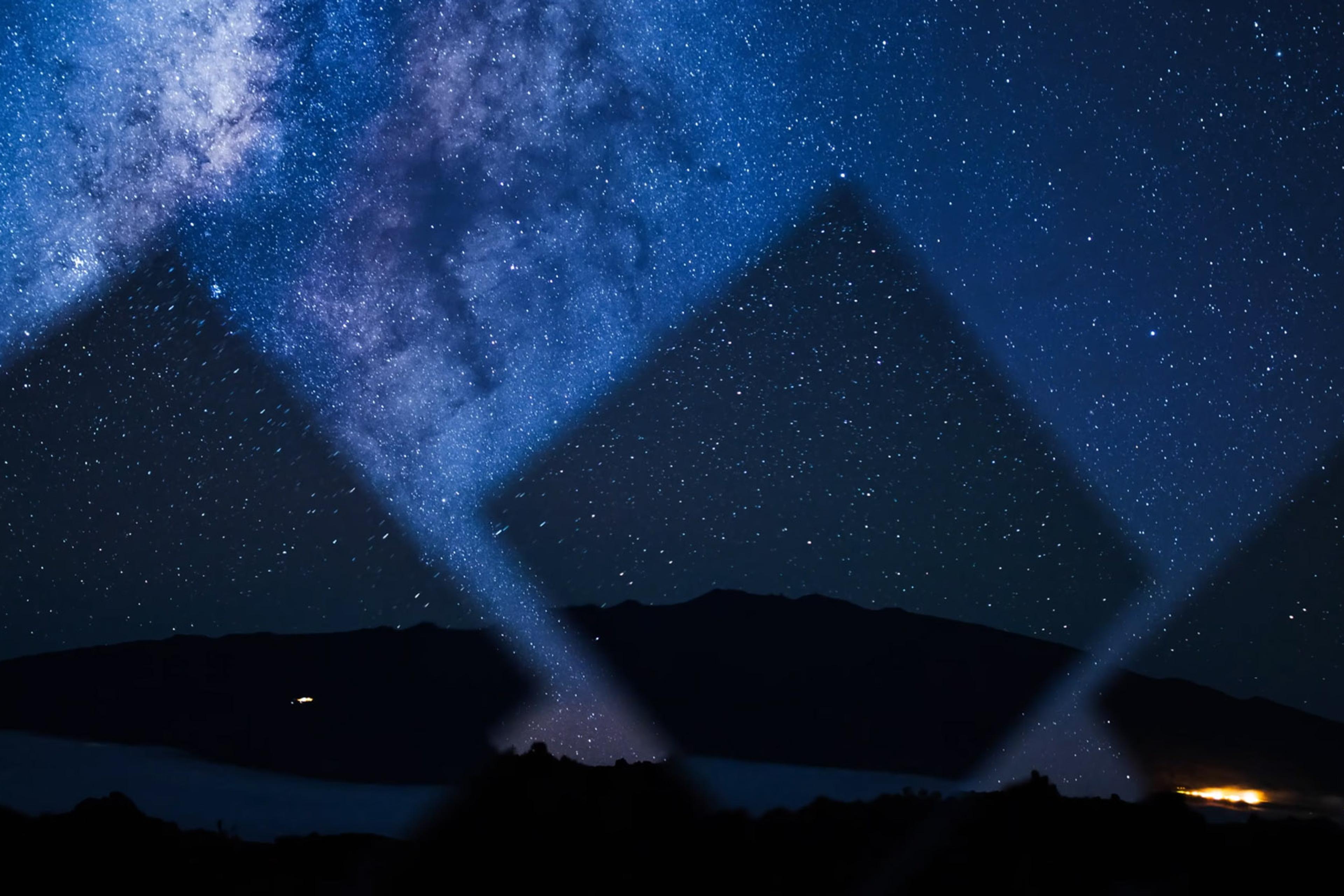 The Milky Way galaxy over a mountainous silhouette at night, with bright spots resembling searchlights crossing the sky.