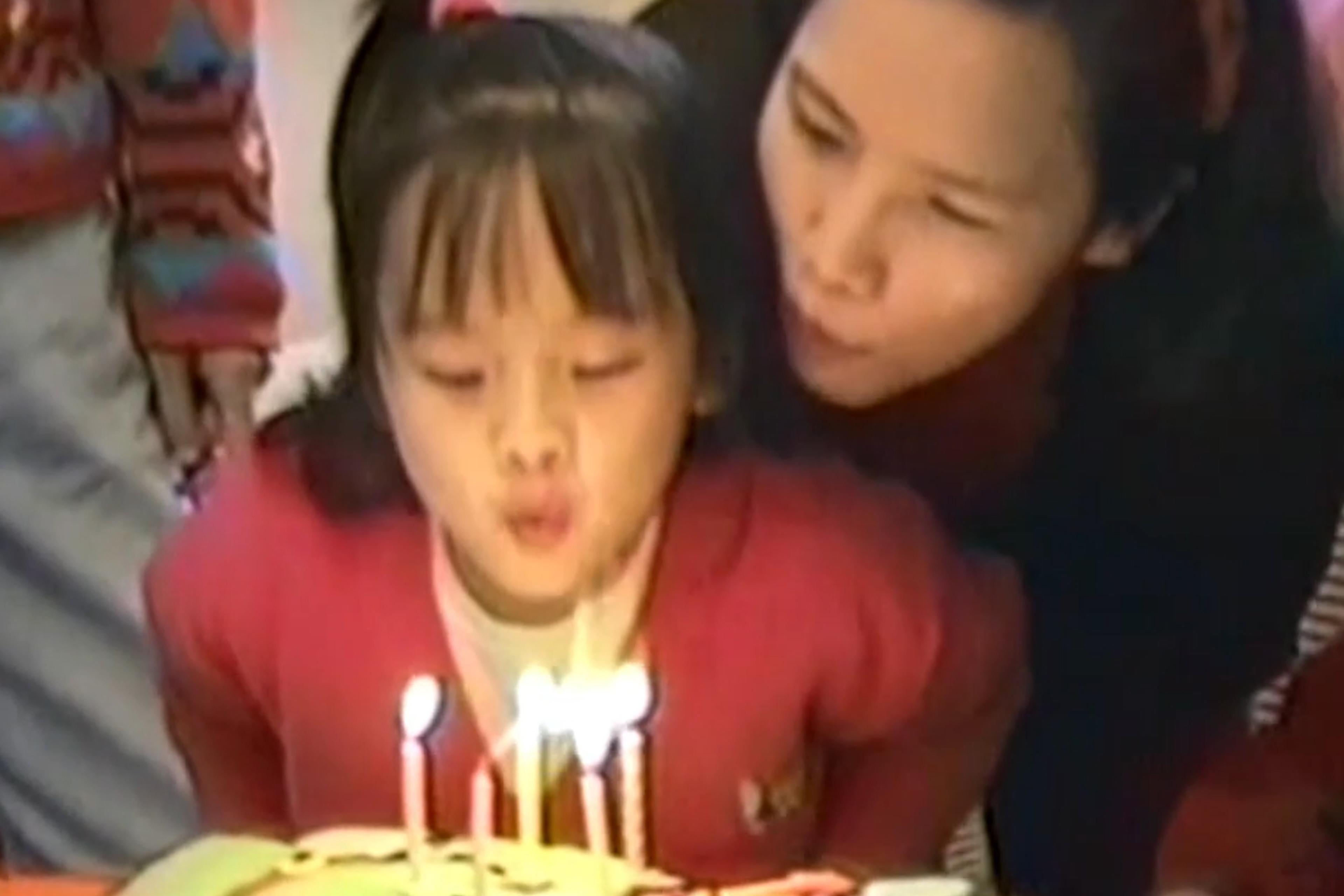 Still image from a VHS tape depicting child and a woman blowing out birthday cake candles together, both wearing red, with people in the background.