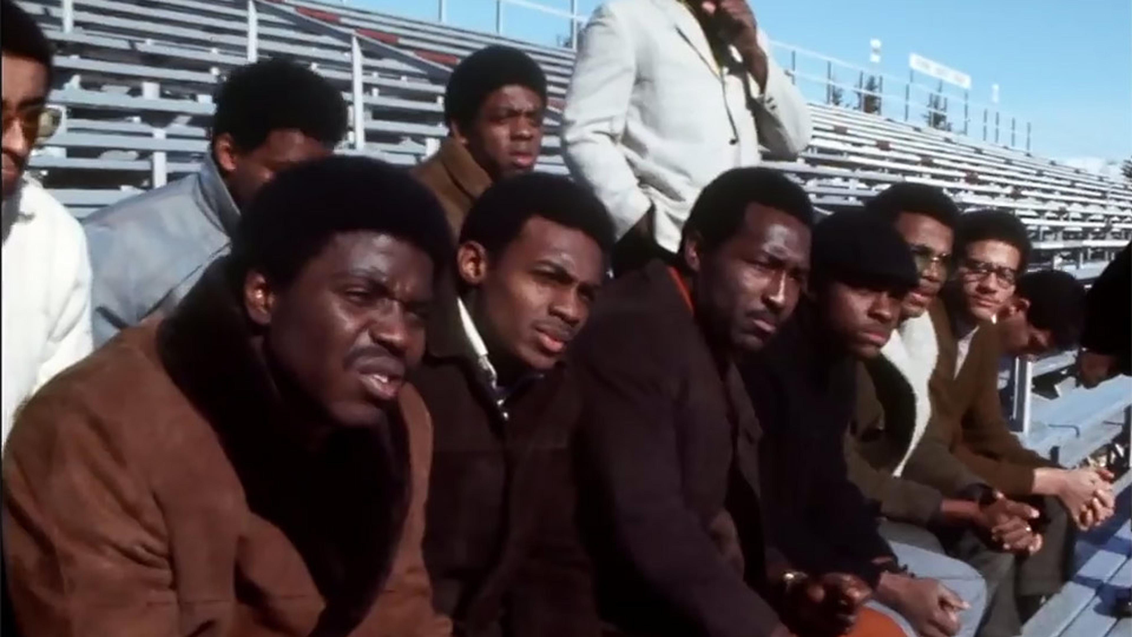 A group of Black men sitting in an outdoor stadium; benches are empty in the background.