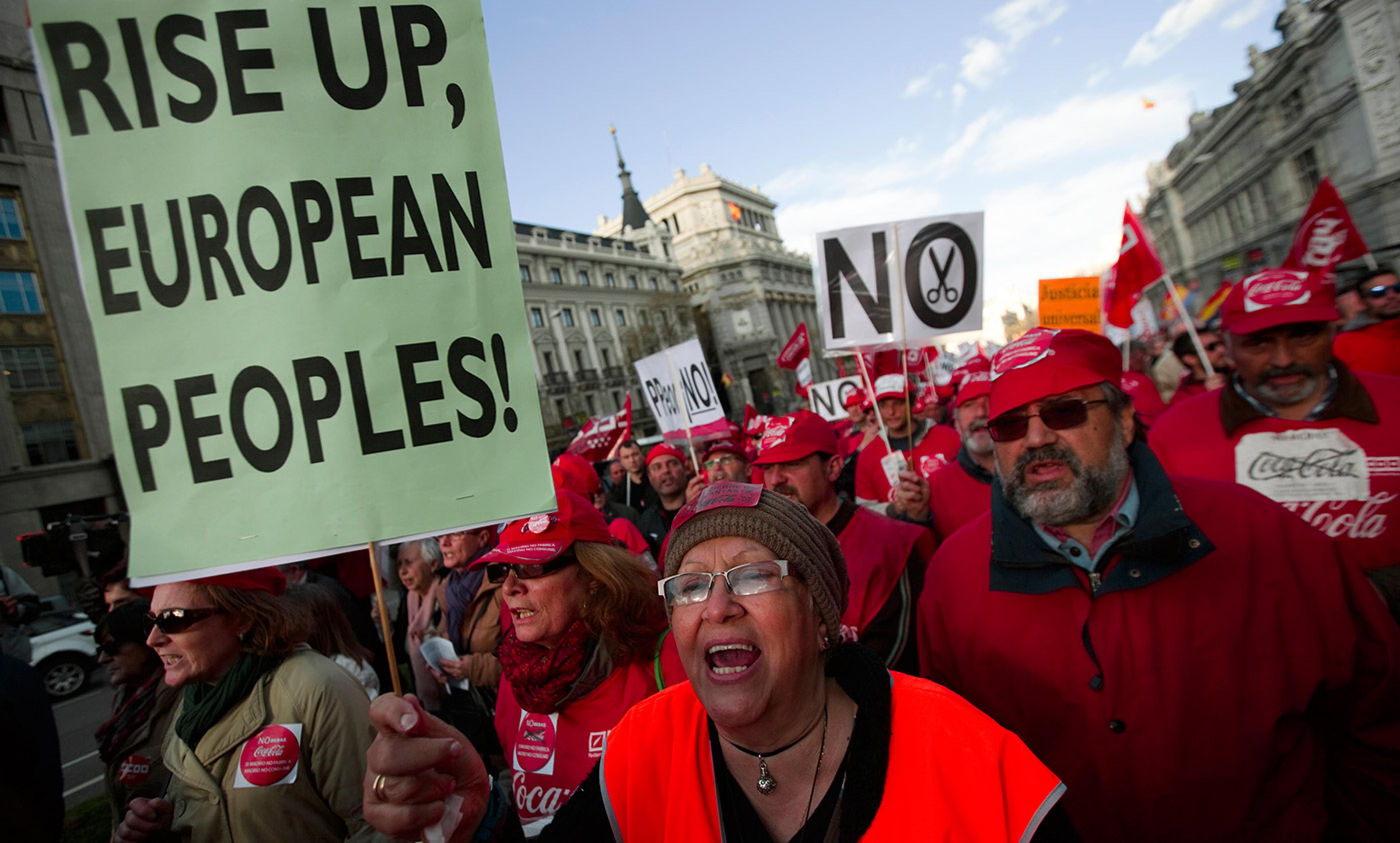 Protesters in a city holding signs, one reads “RISE UP, EUROPEAN PEOPLES!” and another has scissors crossed out.