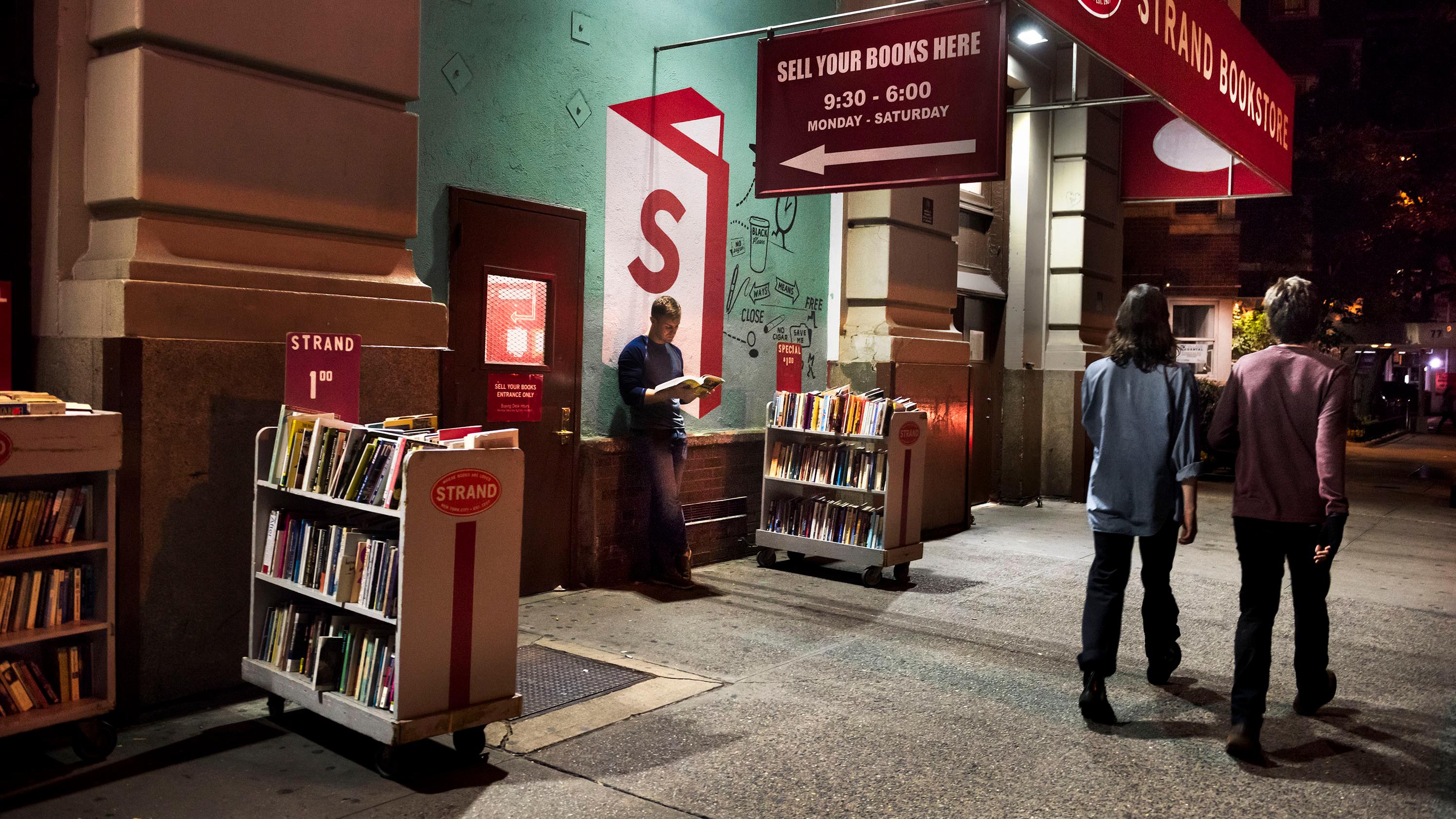 Photo of a bookstore entrance at night with people walking by and a person reading among book trolleys on the pavement.