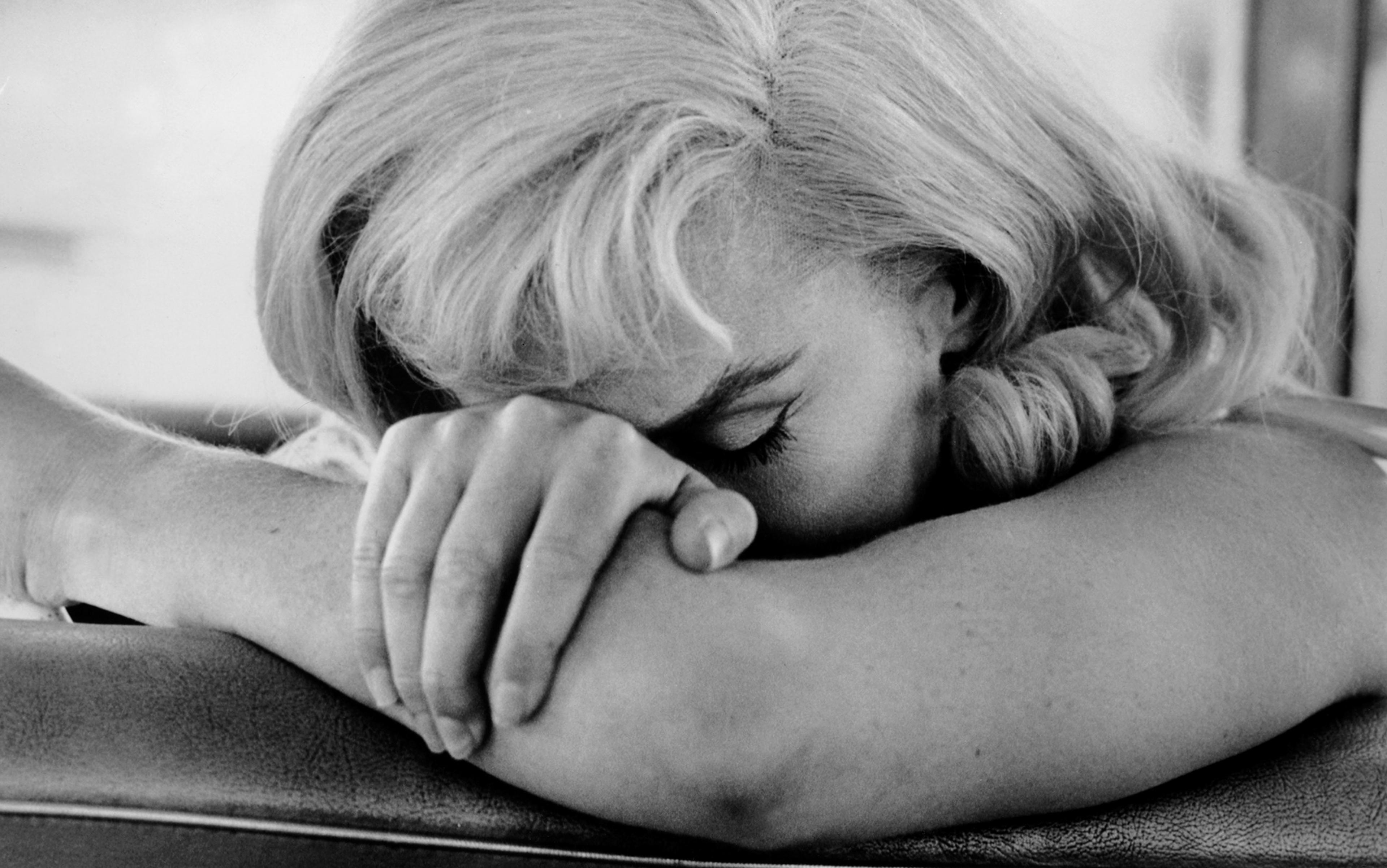 Black-and-white film still of Marilyn Monroe resting her head on her folded arms, her eyes closed, appearing contemplative.