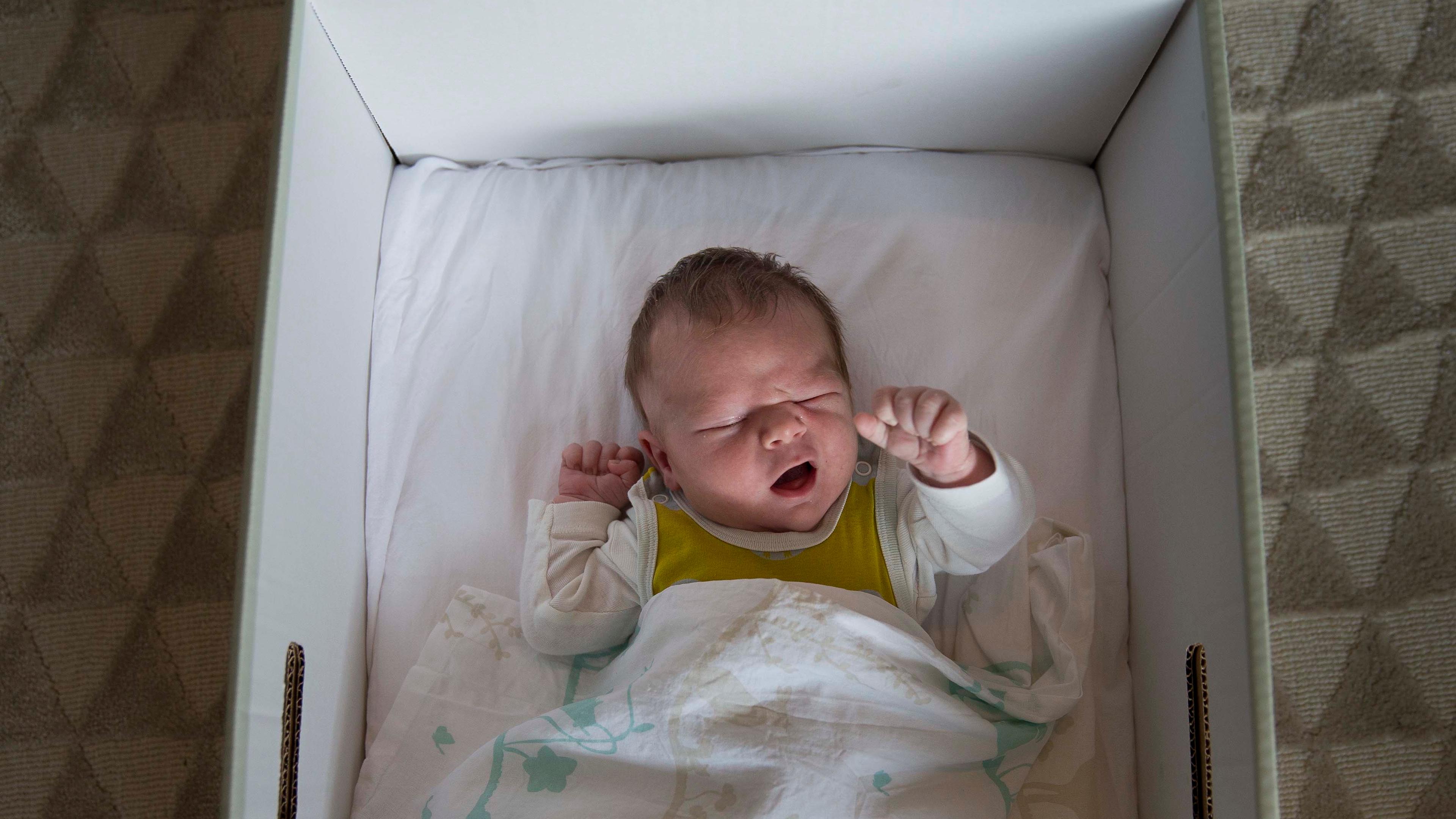 Photo of a newborn baby sleeping in a white box with a blanket on a patterned carpet background.