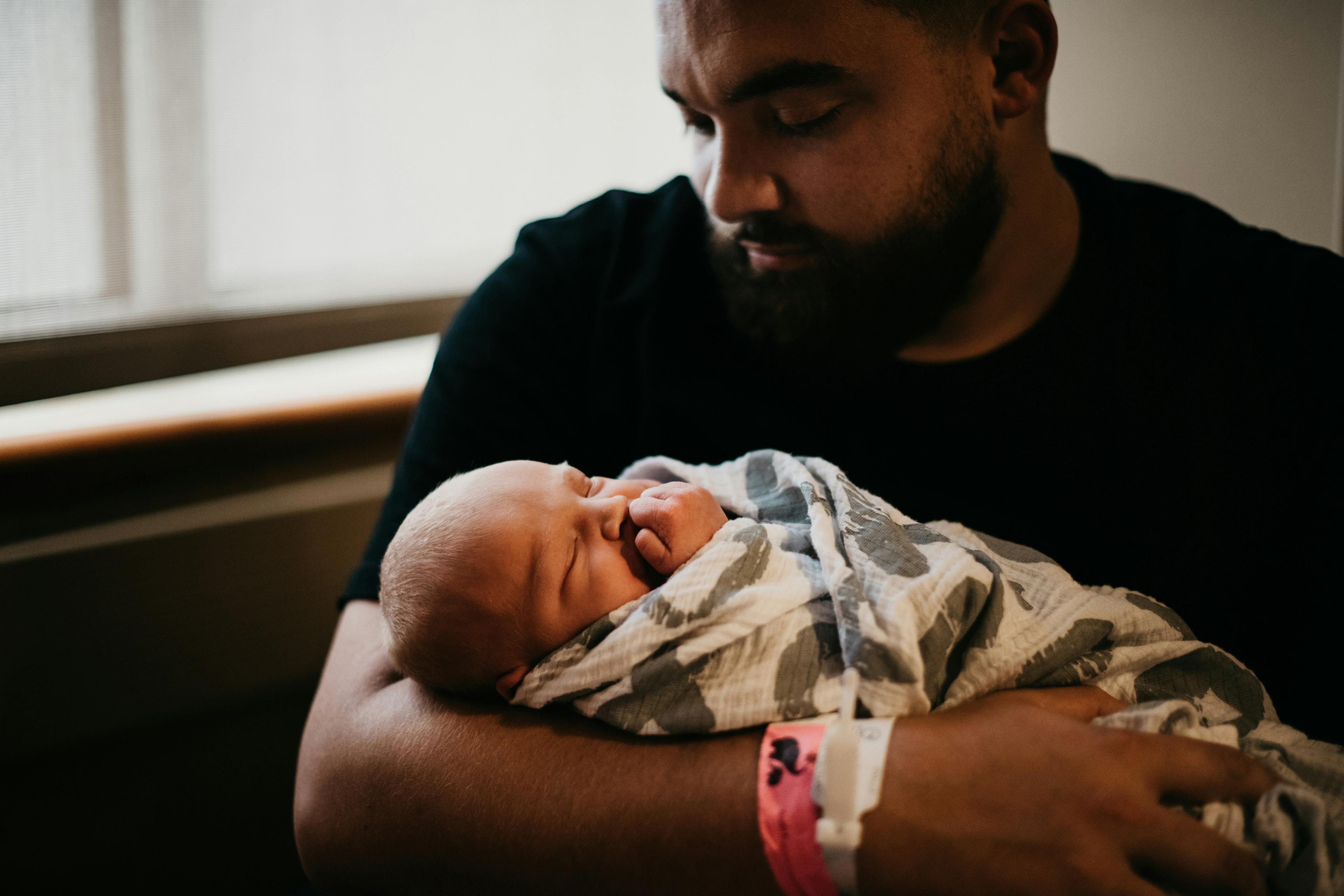 A bearded person gently cradling a newborn wrapped in a patterned blanket by a window in soft light.