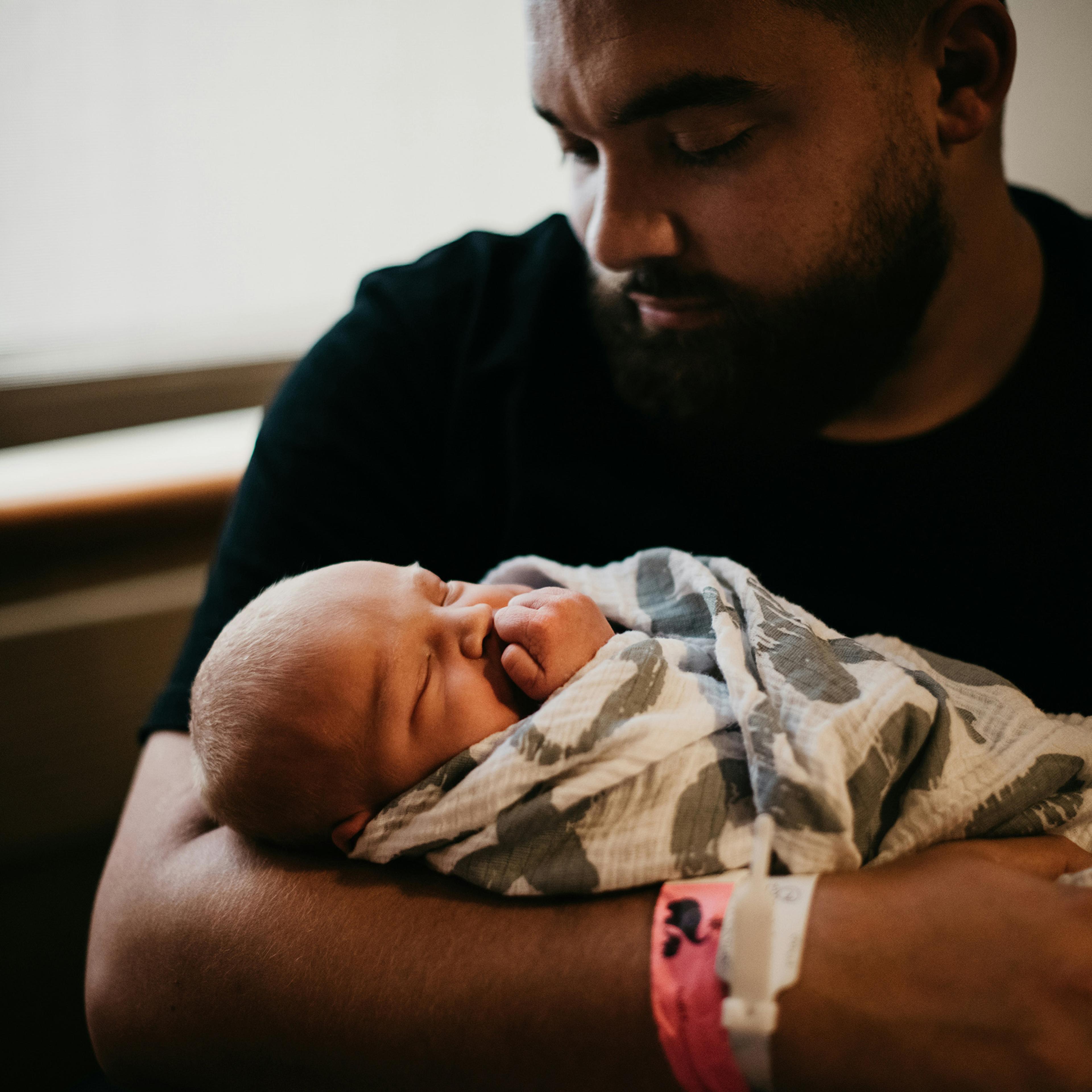 A bearded person gently cradling a newborn wrapped in a patterned blanket by a window in soft light.