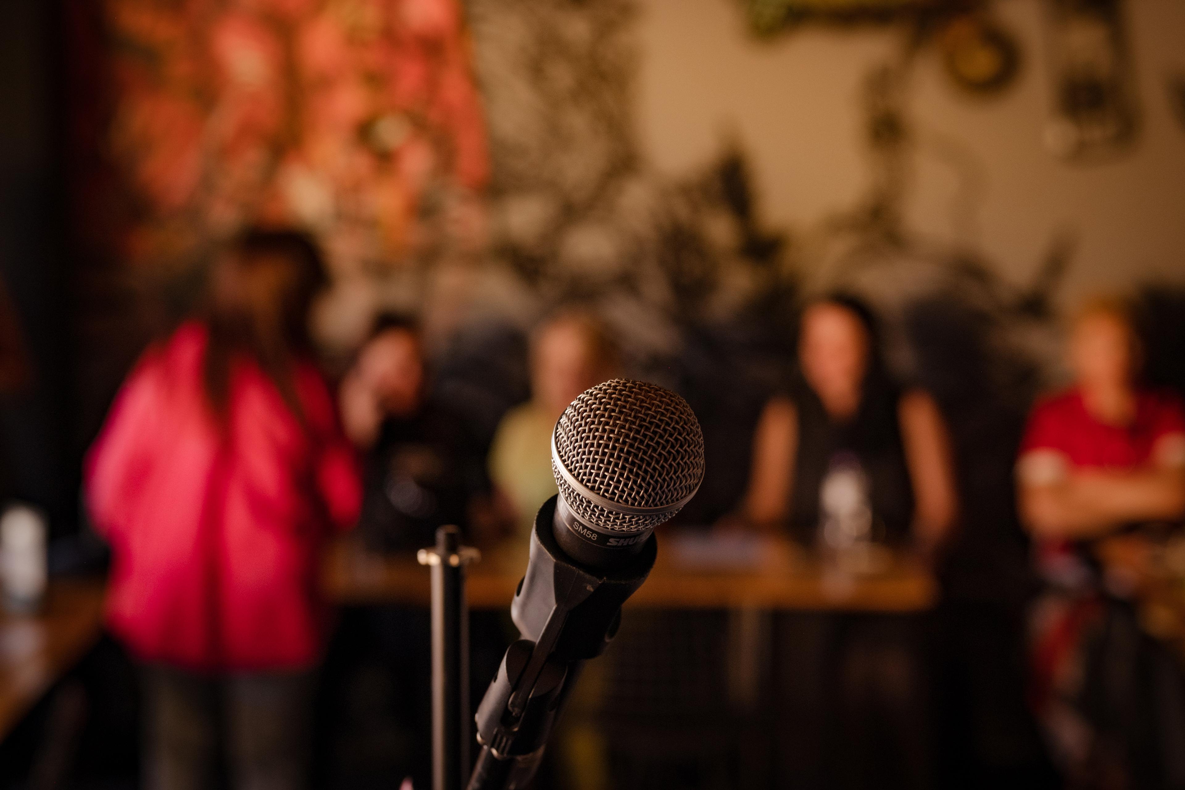 A microphone in focus with a blurred background of people seated at tables in a dimly lit room.