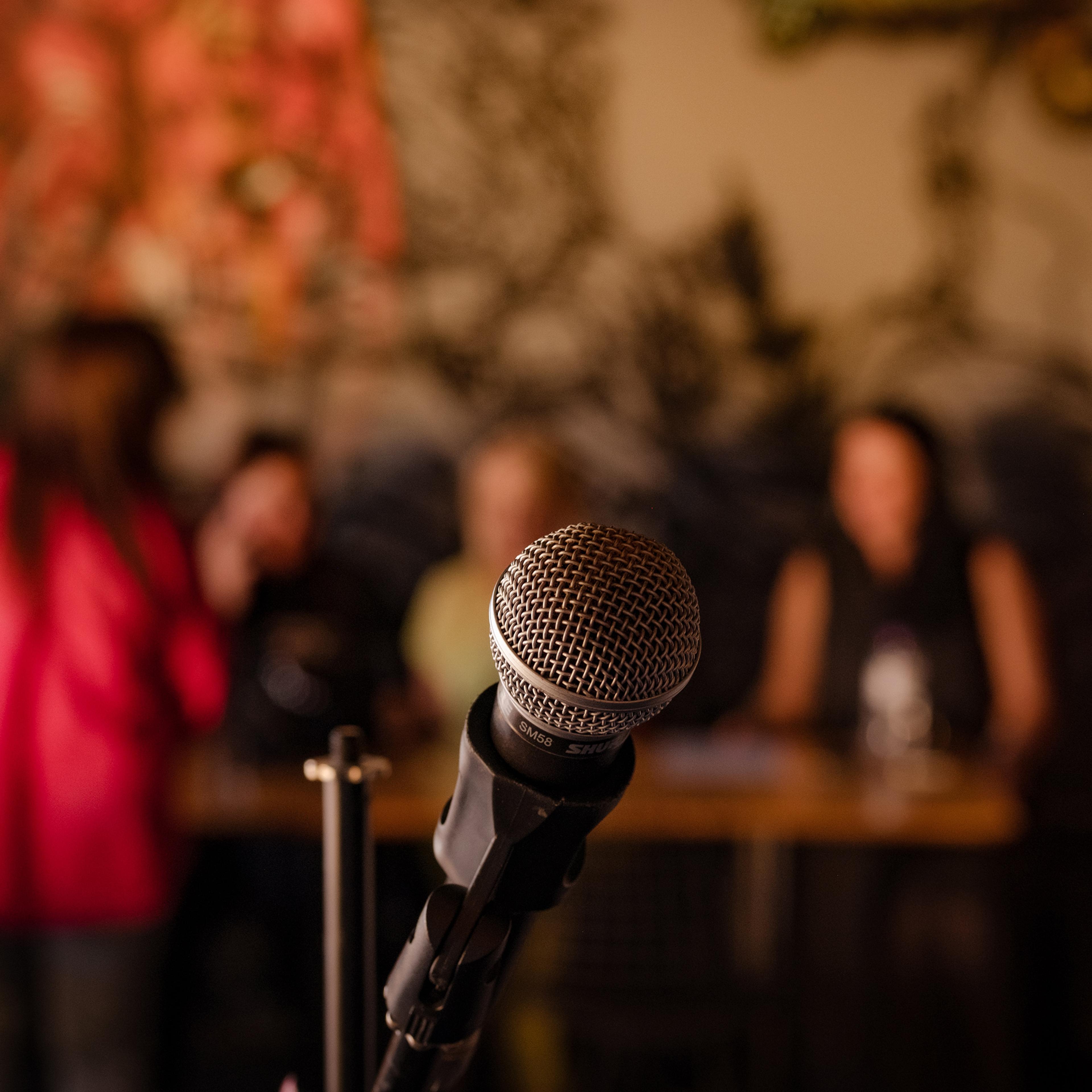 A microphone in focus with a blurred background of people seated at tables in a dimly lit room.