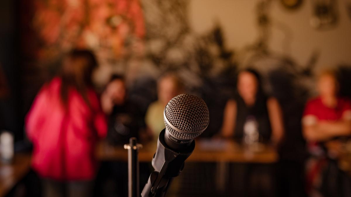 A microphone in focus with a blurred background of people seated at tables in a dimly lit room.