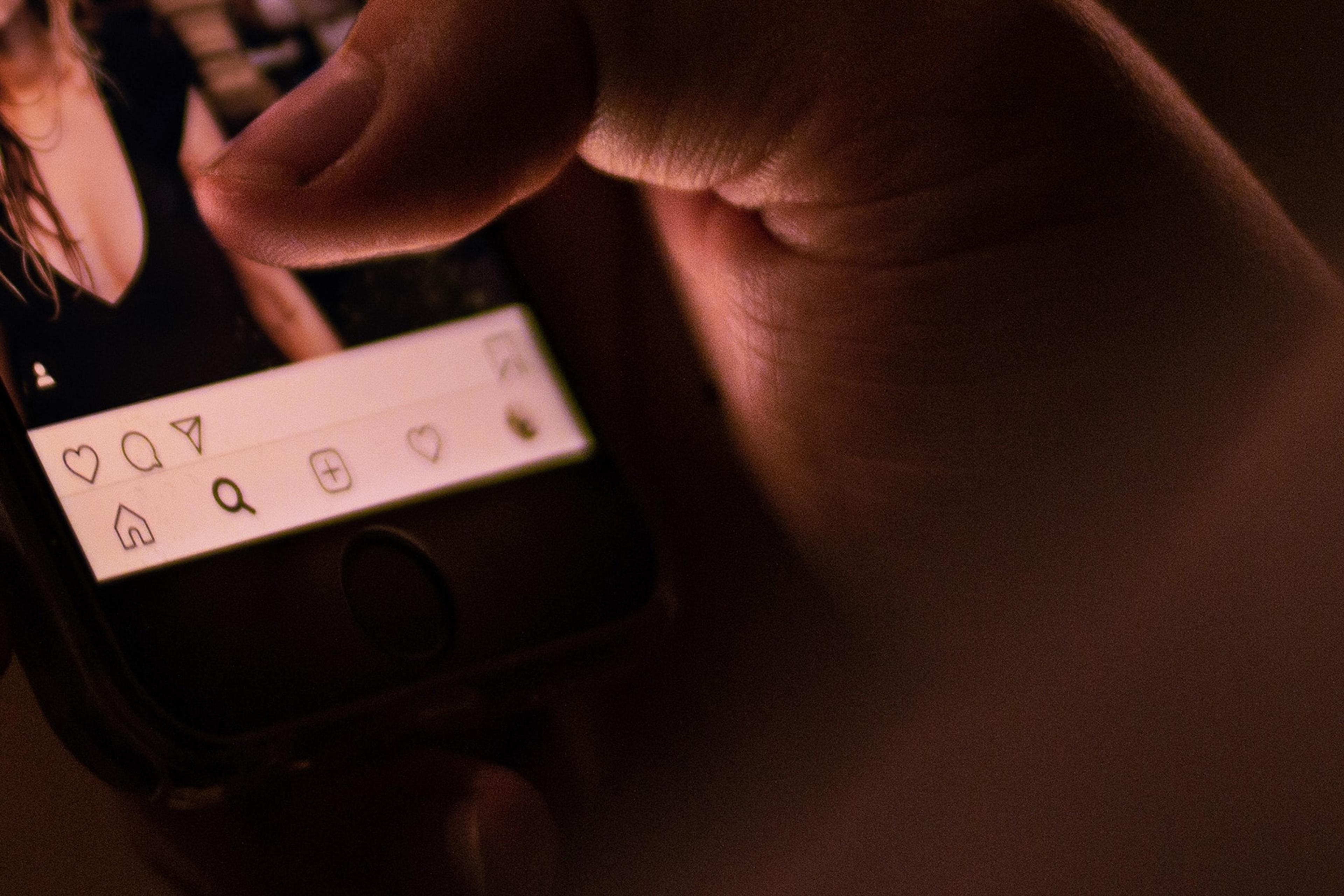 A close-up photo of a hand scrolling through a social media app on a smartphone in dim lighting.