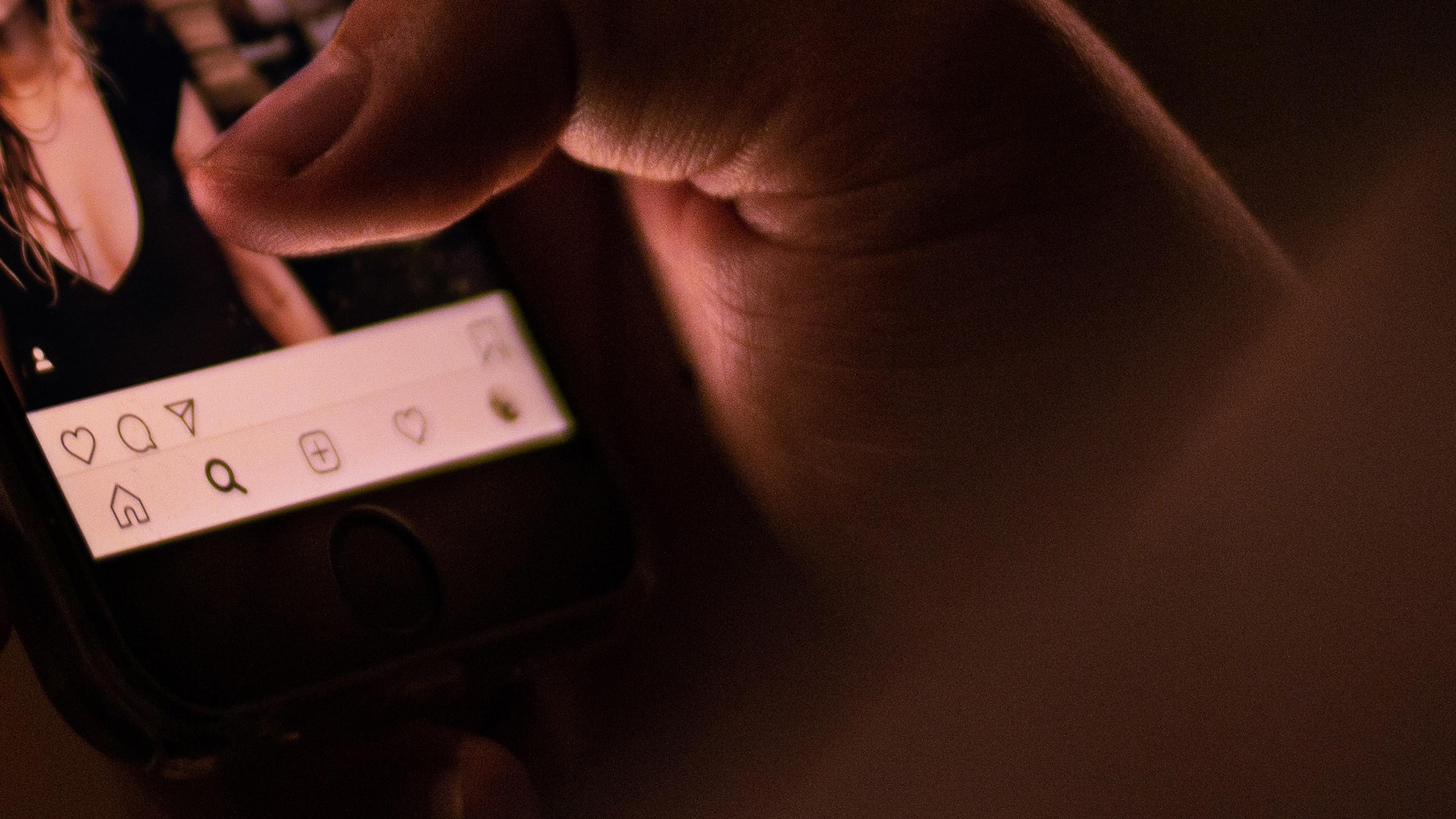 A close-up photo of a hand scrolling through a social media app on a smartphone in dim lighting.