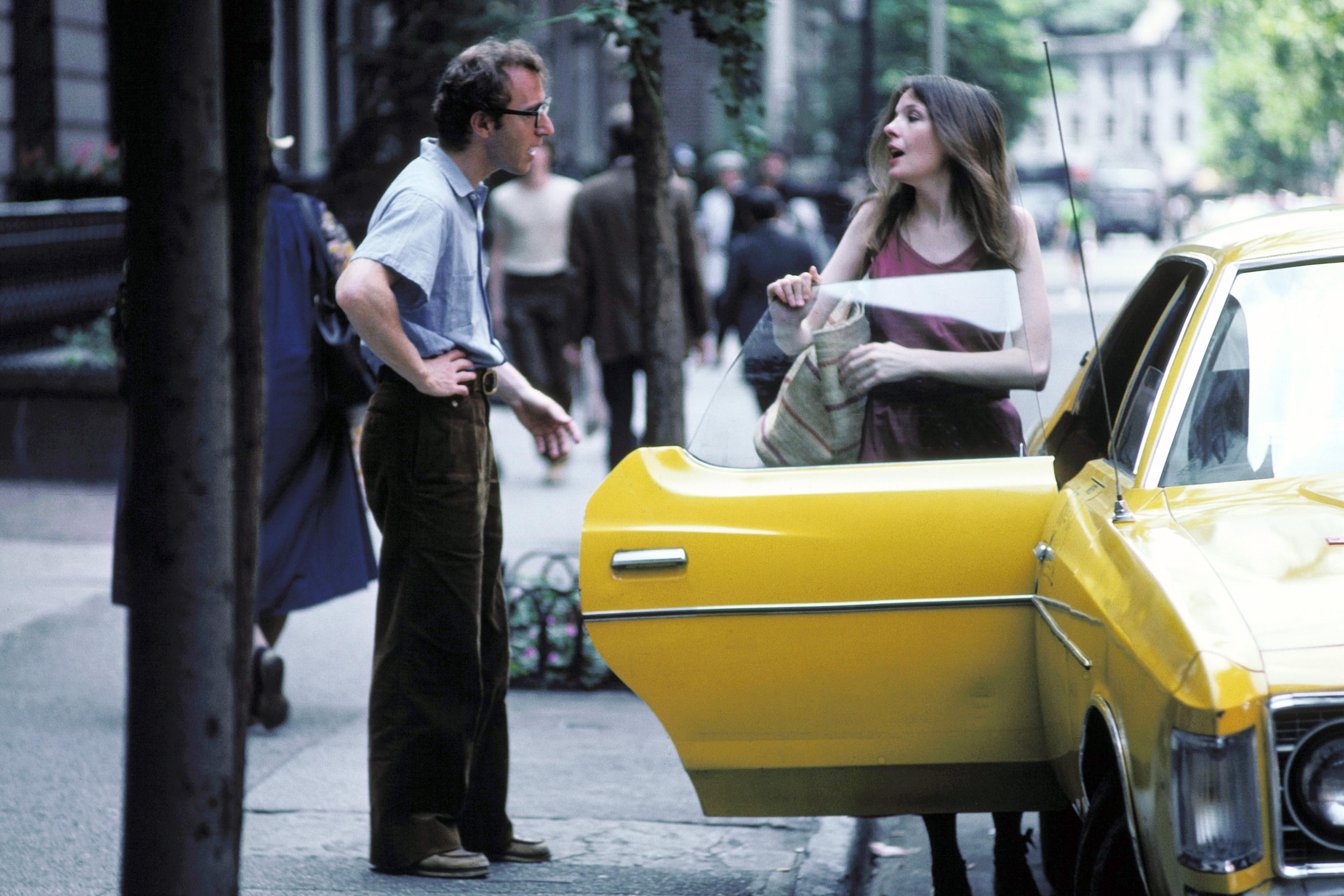 Photo of a man and woman conversing beside a yellow taxi on a city street; the woman is holding the taxi door open.