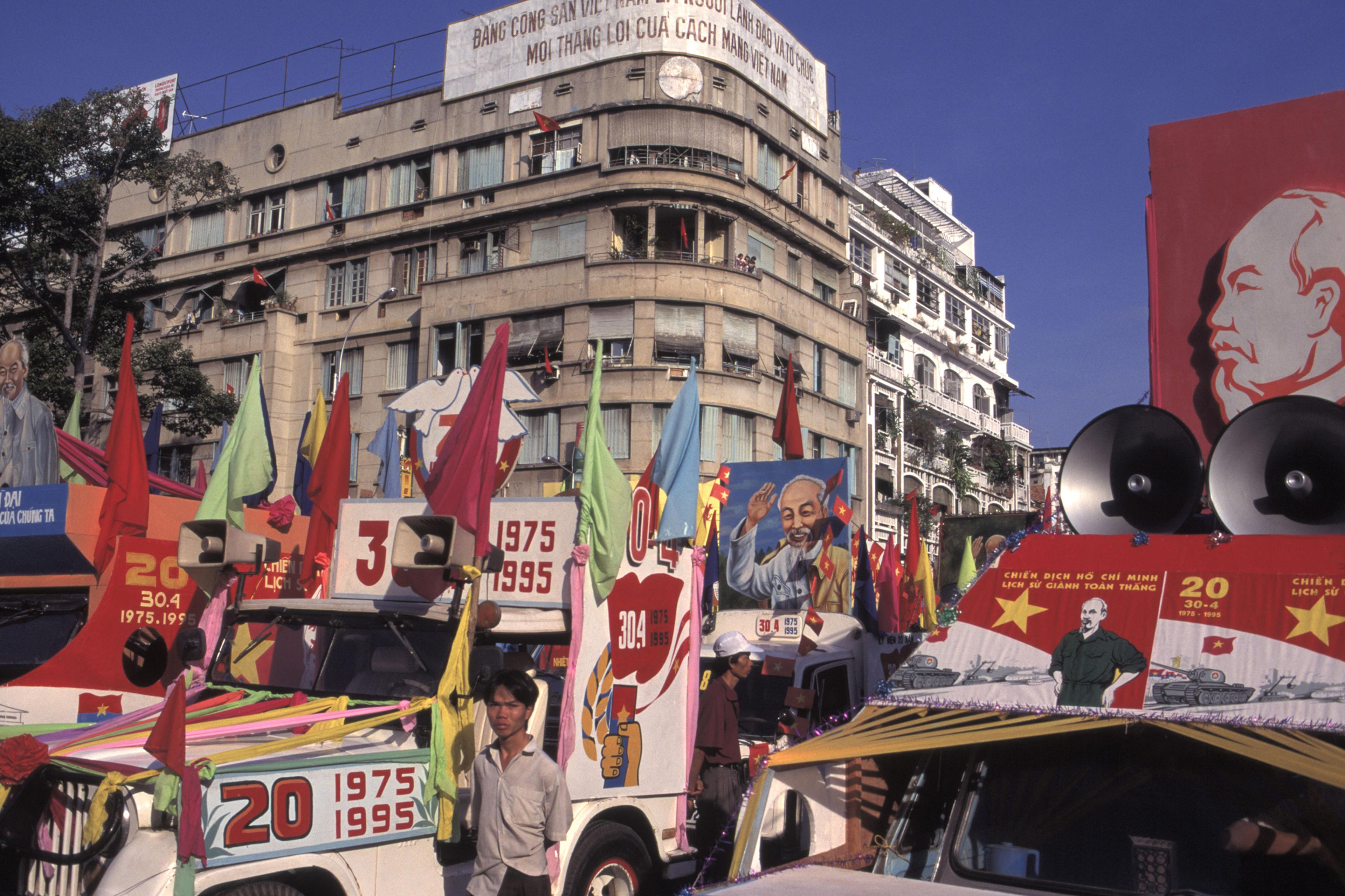 A vibrant street scene with decorated vehicles, colourful flags and banners in front of a large urban building.