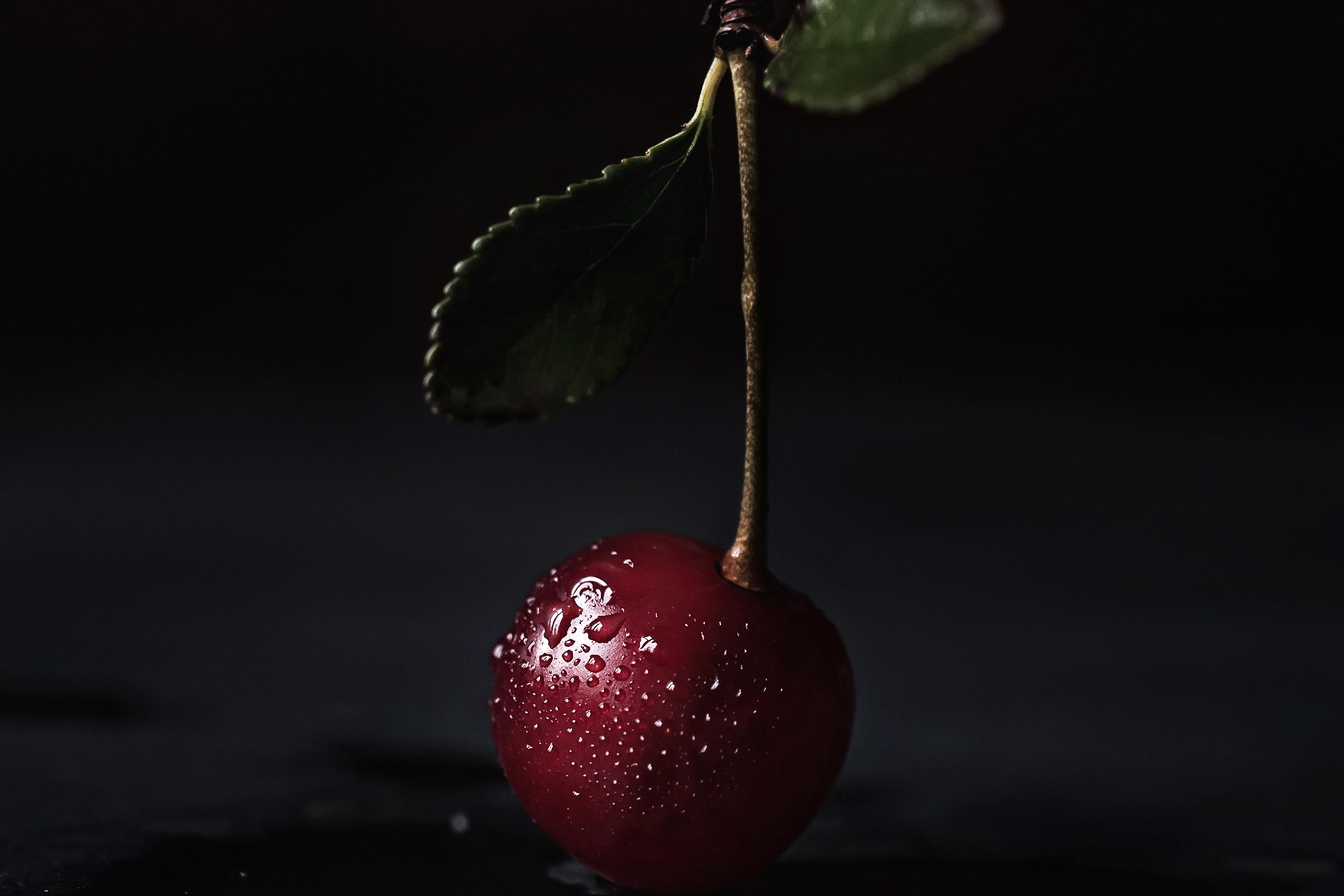 Photo of a single cherry with water droplets and a leaf on a dark background.
