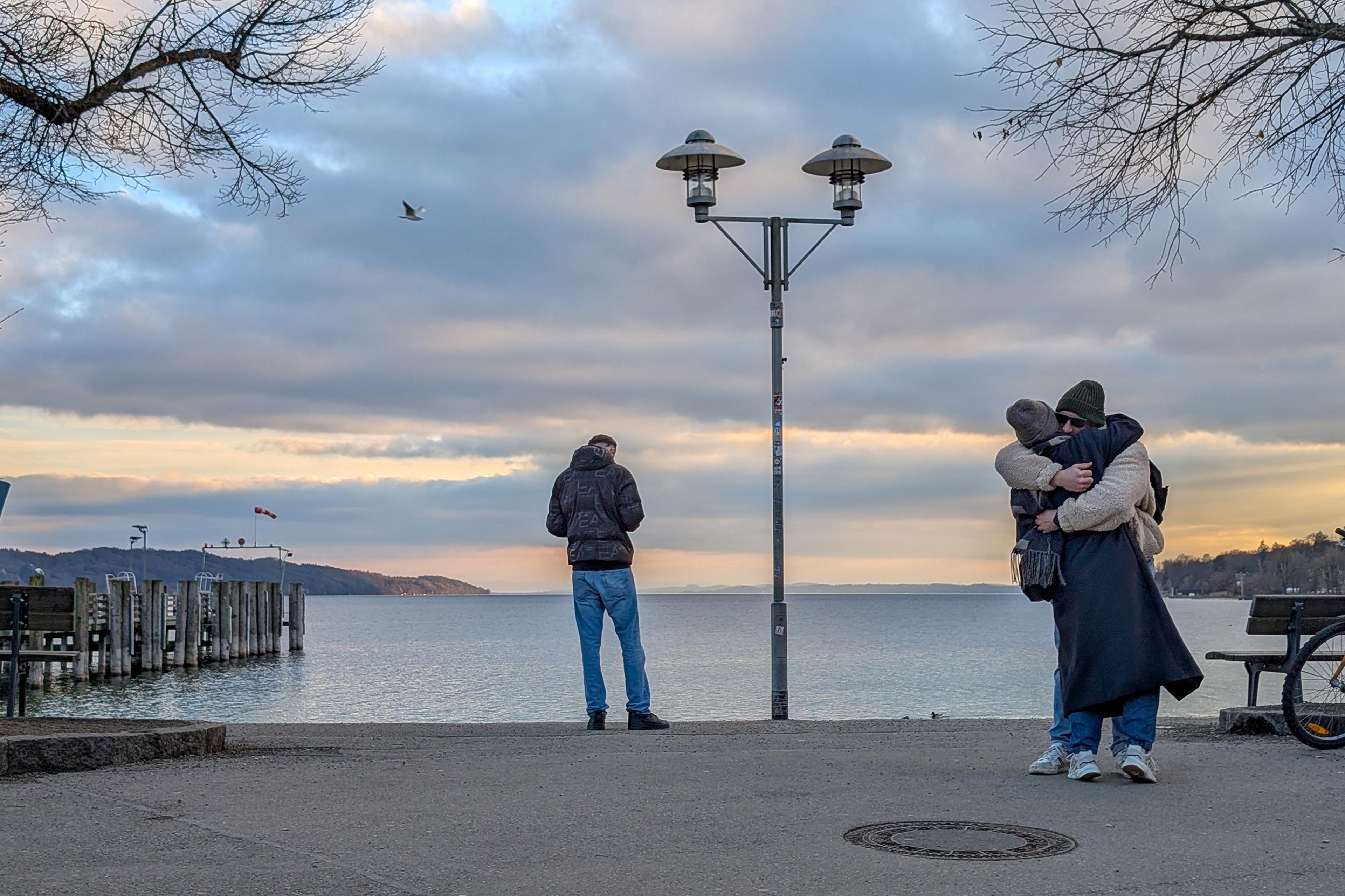 A couple hugging by a lake under a cloudy sky with a person standing by a lamppost nearby.