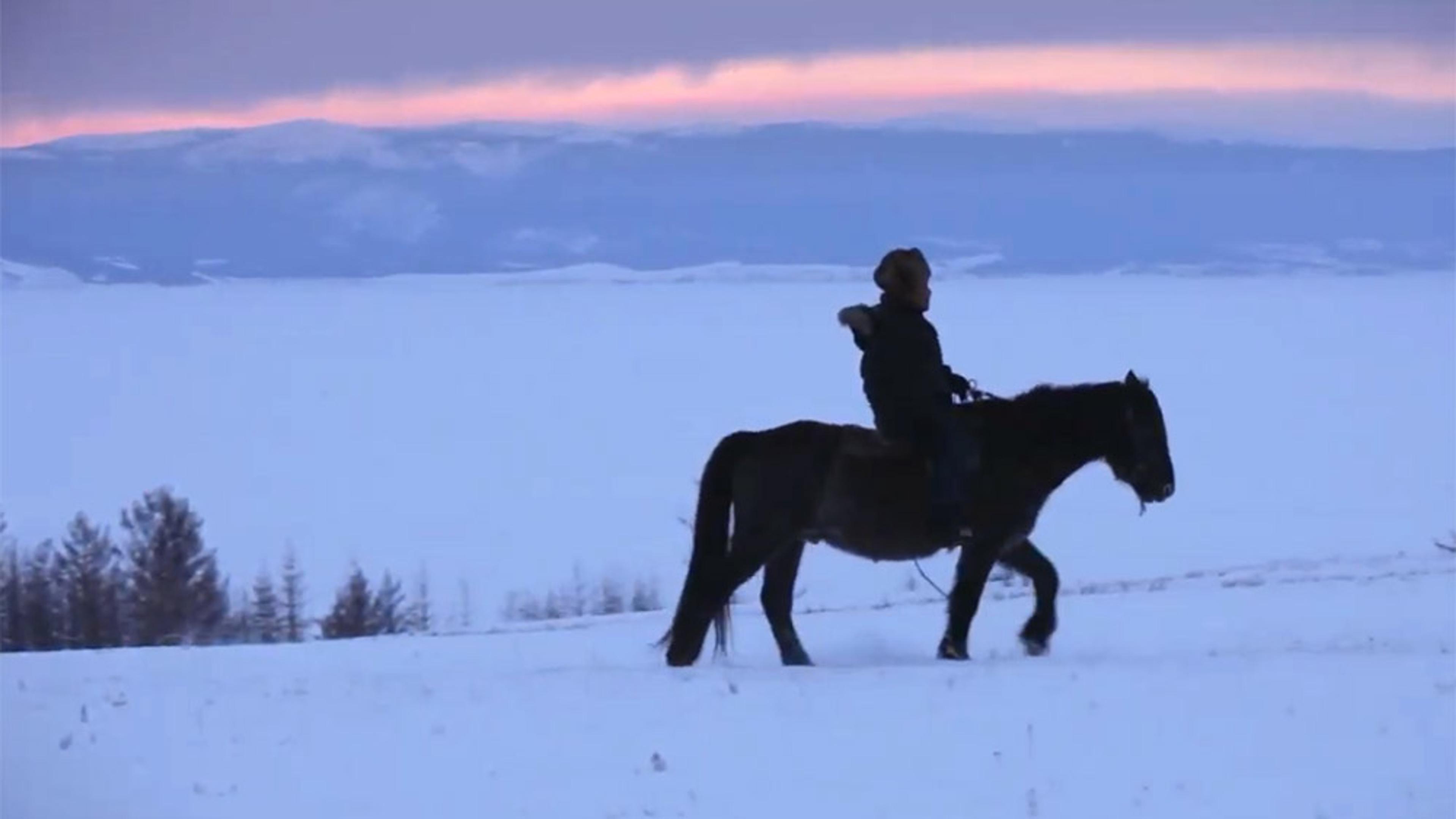 A person riding a horse across a snowy landscape at dusk with a mountain range in the background and pink sky.